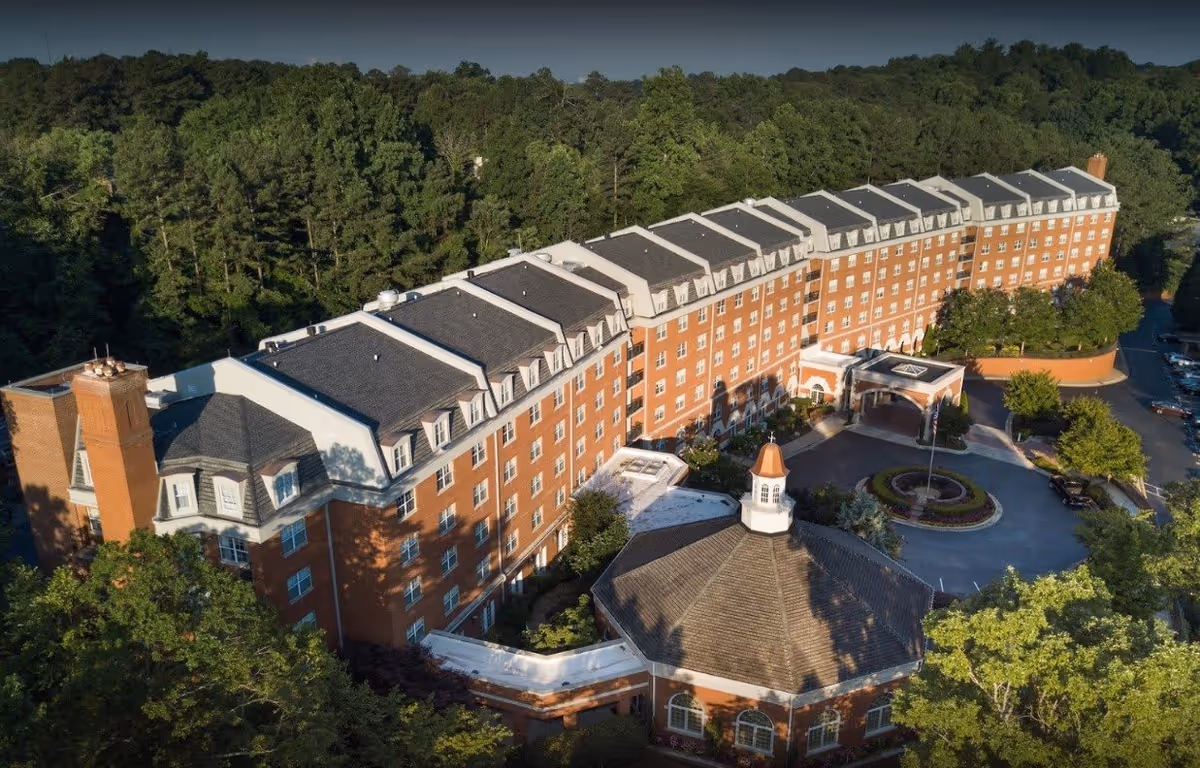 Aerial view of King's Bridge Retirement Community showing a large multi-story brick building with a distinctive octagonal structure in front, surrounded by trees and greenery.