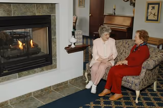 Two elderly women sitting and talking on a floral patterned couch in a cozy living room with a lit fireplace on the left and a piano in the background.