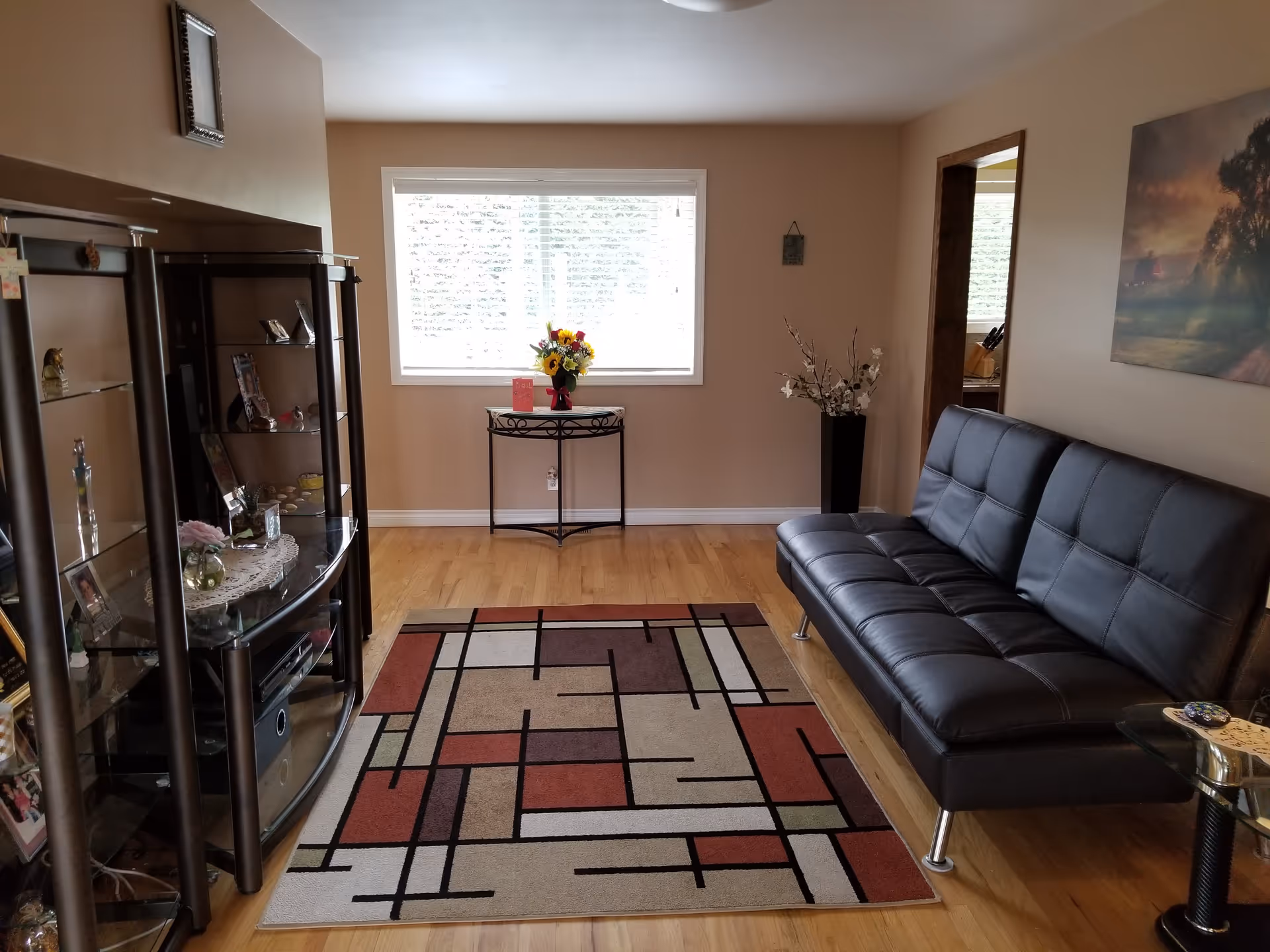A cozy living room with a black leather couch on the right, a colorful geometric patterned rug on a wooden floor, a glass and metal entertainment center on the left with various decorative items, a small table with a flower vase in front of a large window with blinds, and a tall vase with white flowers near the corner.