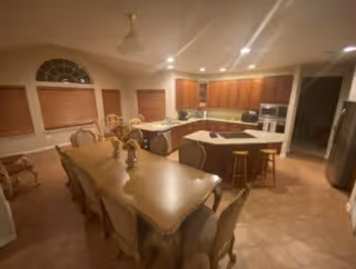 Interior view of a kitchen and dining area in a senior living facility. The dining area features a long table with multiple chairs and decorative items on top. The kitchen has wooden cabinets, a central island with bar stools, and modern appliances including a microwave and oven. The room is warmly lit with recessed lighting and ceiling fans, and windows with closed blinds are visible.