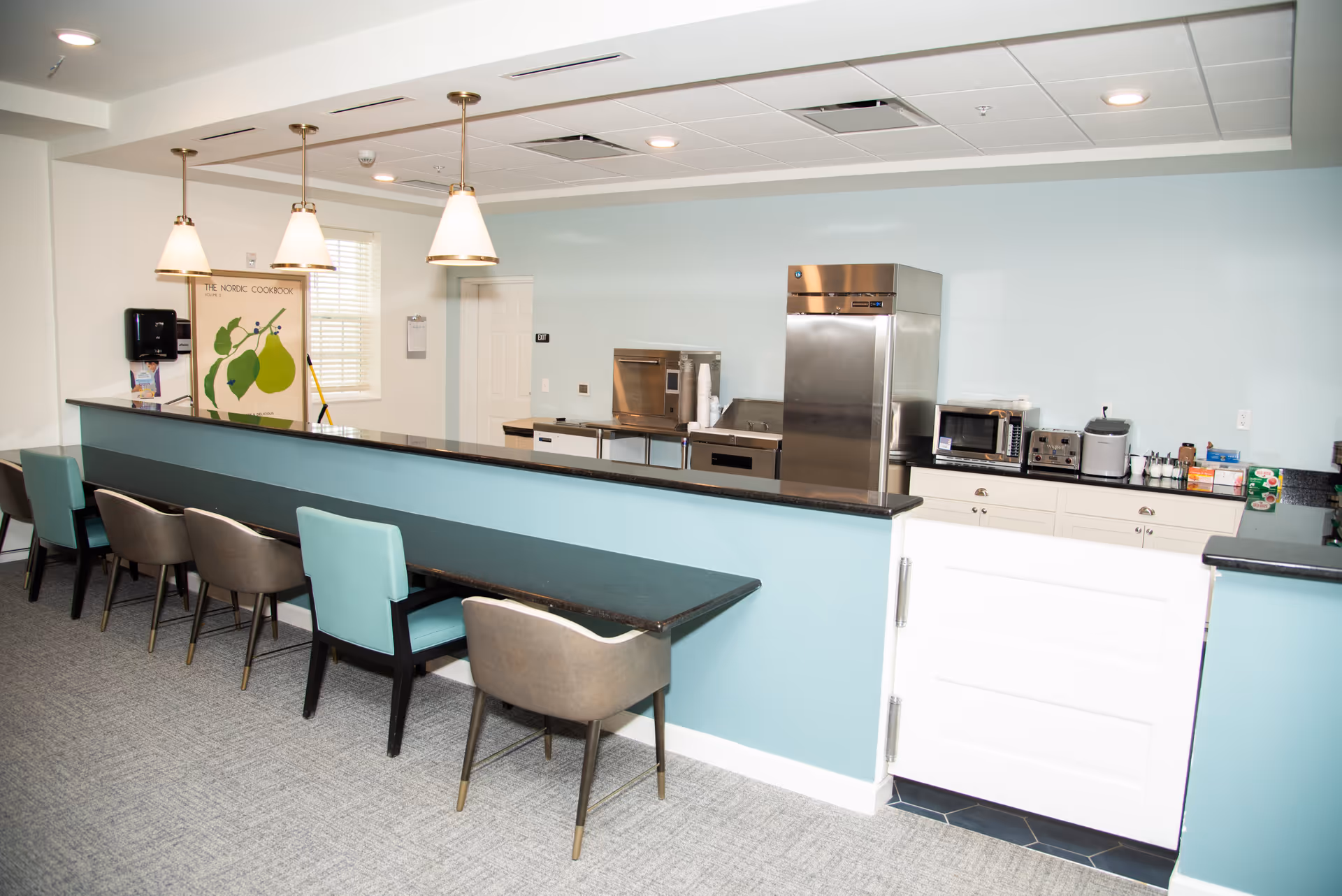 Interior view of a kitchen area in a senior living facility with a long counter and several chairs lined up in front. The kitchen has stainless steel appliances including a refrigerator, microwave, and toaster, with cabinets and a light blue wall. Three pendant lights hang from the ceiling above the counter.