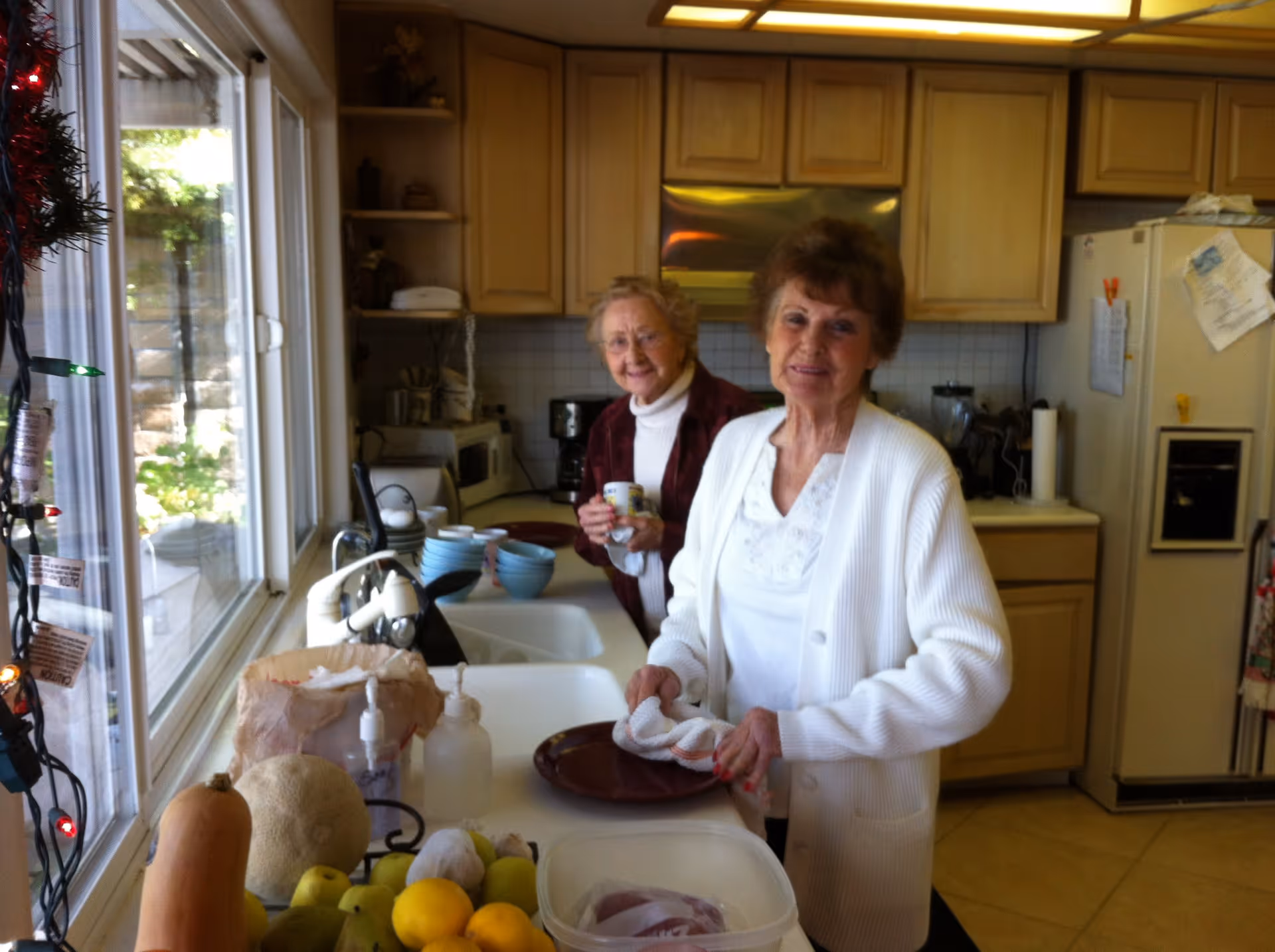 Two elderly women standing in a kitchen. One woman in a white cardigan is drying a plate with a towel near the sink, while the other woman in a maroon jacket is holding a cup and smiling. The kitchen has light wood cabinets, a refrigerator with papers attached, a microwave, and various kitchen items on the counter. A window to the left shows some greenery outside.