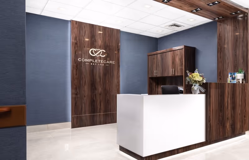 Reception area of Complete Care at Bey Lea featuring a modern white reception desk with dark wood paneling behind it, a matching cabinet, a flower arrangement on the desk, and the facility's logo on the wood-paneled wall.
