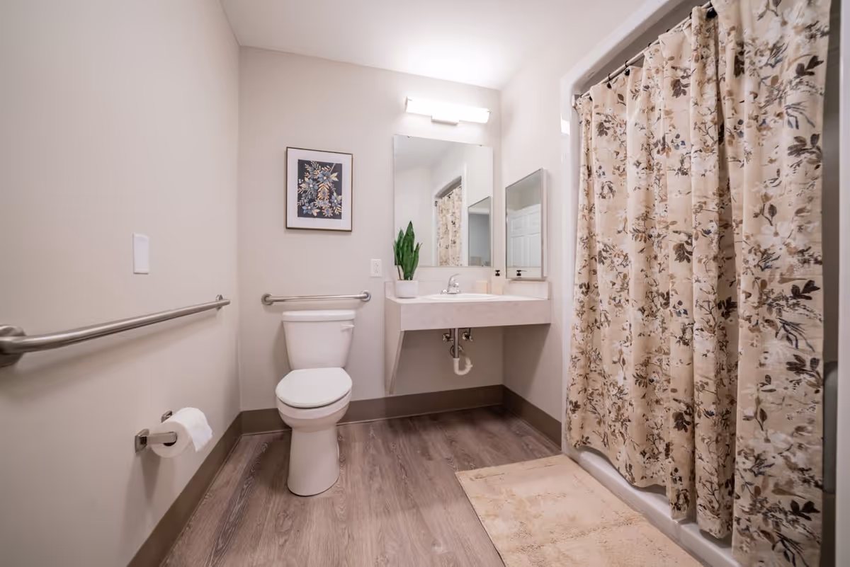 Accessible bathroom with a toilet, wall-mounted sink and mirror, grab bars, and a floral shower curtain.