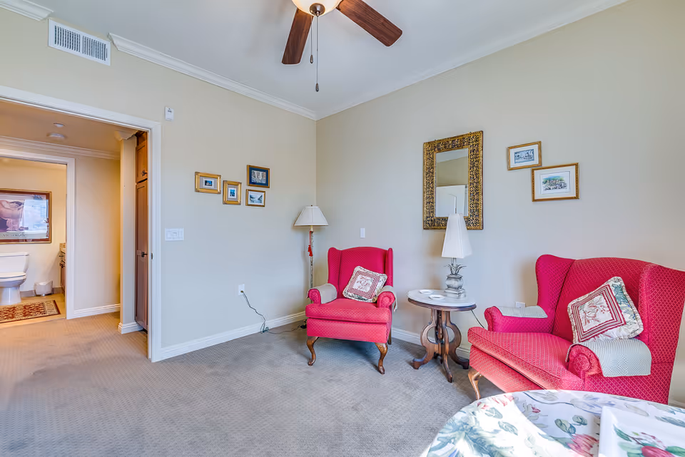 Cozy sitting area with two red upholstered armchairs flanking a small round table and lamp, a mirror on the wall, and a doorway leading to a bathroom.