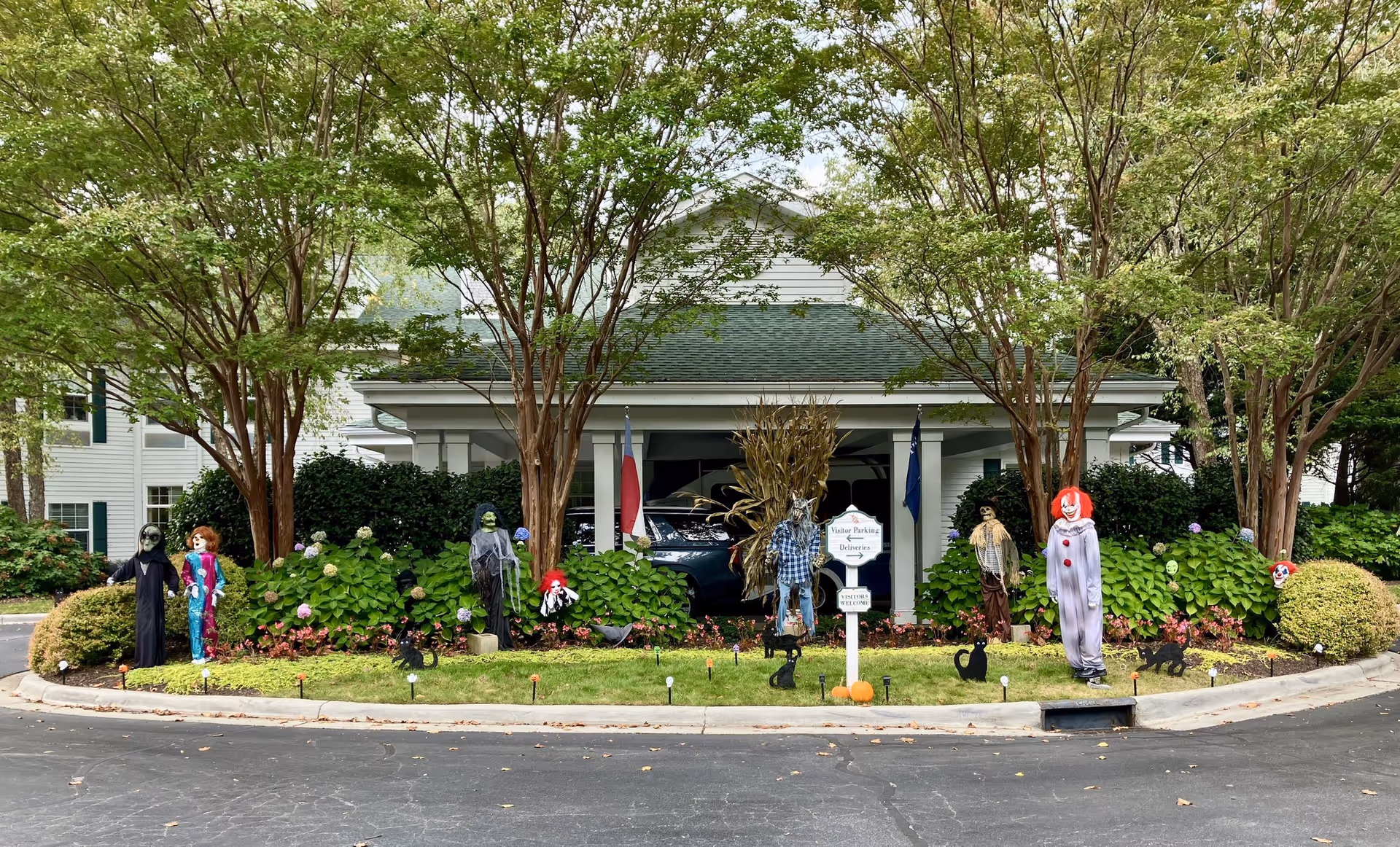 Front exterior view of Arbor Terrace Asheville decorated with Halloween-themed figures including clowns, scarecrows, and other spooky characters among green bushes and trees.