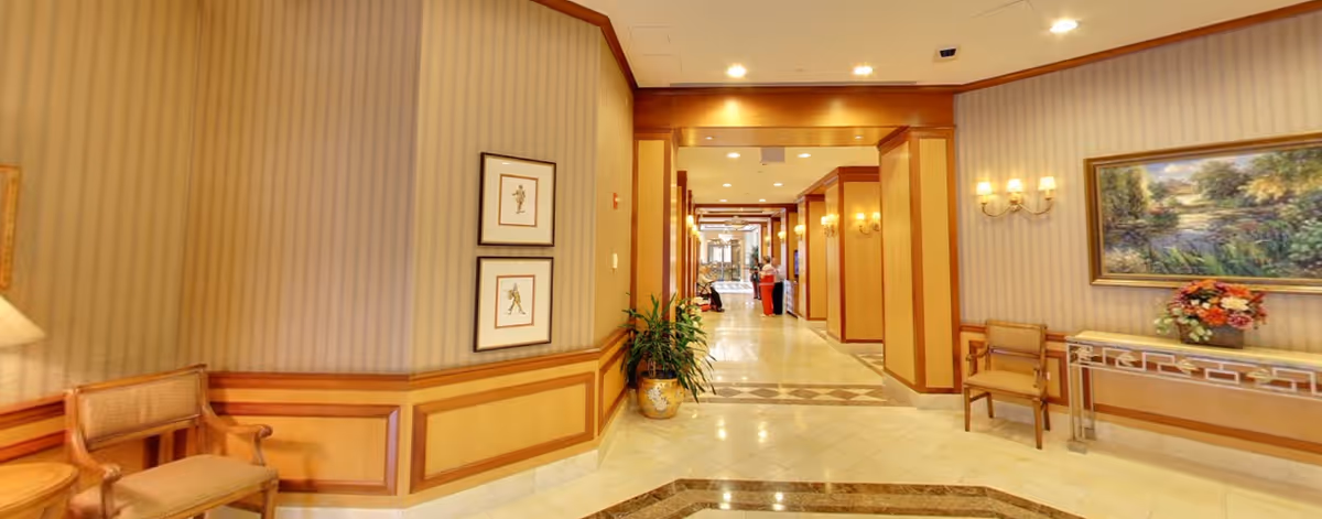 A well-lit hallway in a senior living facility with beige striped wallpaper and wooden paneling. The hallway features framed artwork on the walls, a potted plant, a console table with a floral arrangement, and several chairs along the walls. Two people are visible in the distance near the end of the hallway.