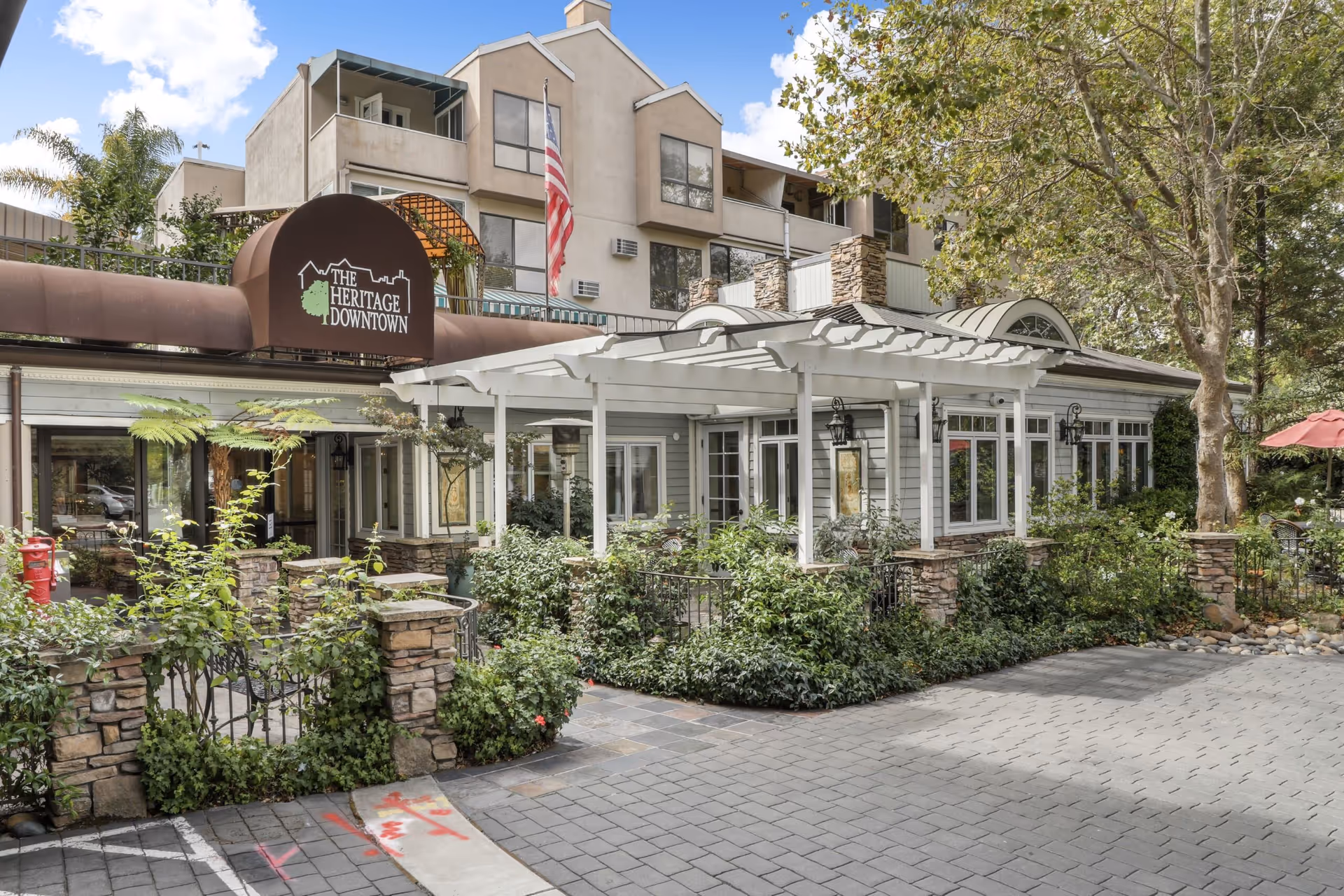 Exterior view of The Heritage Downtown senior living facility showing a multi-story building with beige walls and multiple windows. In front, there is a white pergola over a garden area with various green plants and stone pillars. An American flag is visible near the entrance under a brown awning with the facility's name and logo.