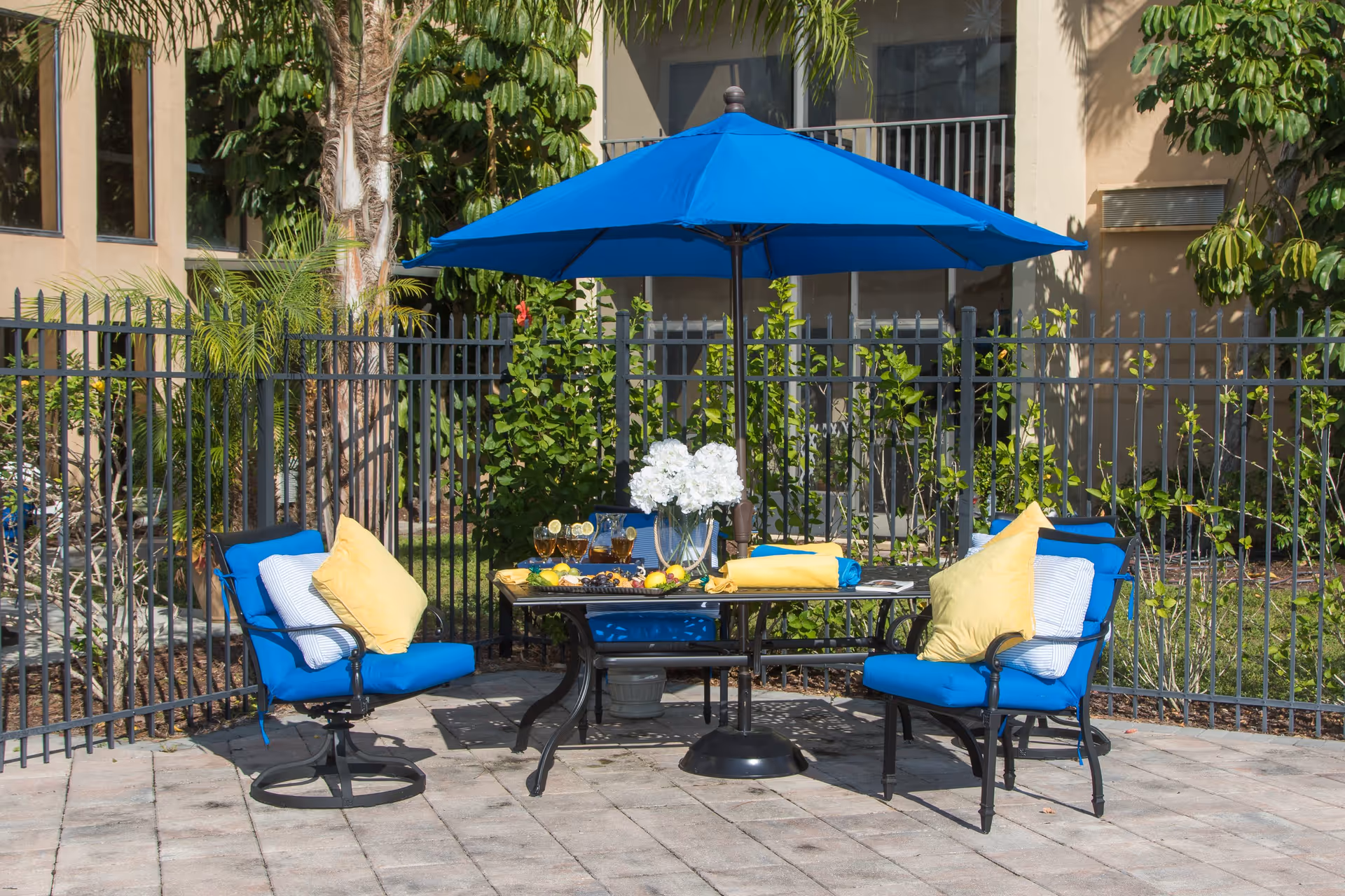 Outdoor patio area with a black metal table and four blue cushioned chairs with yellow and white pillows. A large blue umbrella shades the table, which is set with glasses of iced tea, a pitcher, a plate of fruit, and a vase of white flowers. The area is enclosed by a black metal fence with green plants and a building in the background.