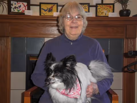 An elderly woman with glasses and gray hair wearing a purple sweater is sitting in a chair in front of a fireplace. She is holding a small black and white dog wearing a pink bandana. Behind her on the mantel are several framed colorful artworks and a potted plant.