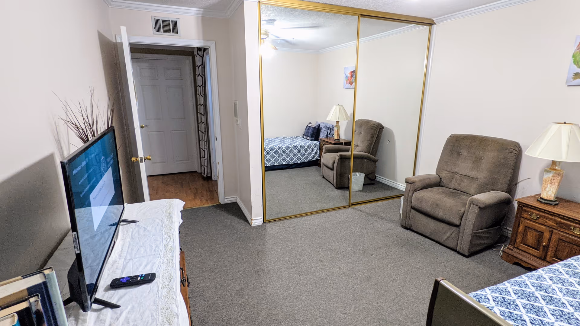 A cozy assisted living bedroom with a patterned blue and white bedspread, a brown recliner chair, a wooden nightstand with a lamp, and a large mirrored closet door reflecting the room. A TV is placed on a dresser opposite the bed, and an open door leads to a hallway with wooden flooring.