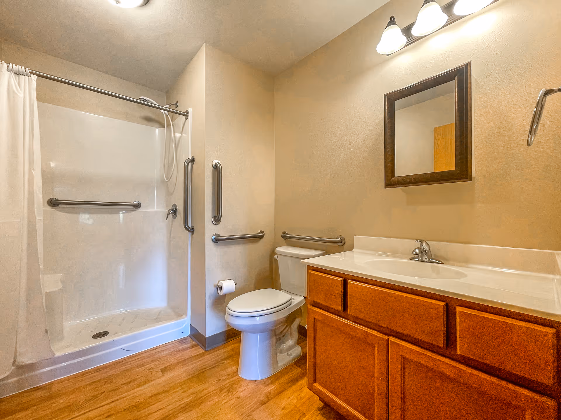 A bathroom with a walk-in shower featuring grab bars and a white curtain, a toilet with grab bars on the walls nearby, and a wooden vanity with a sink and a mirror above it. The floor has a wood-like finish and the walls are painted beige.