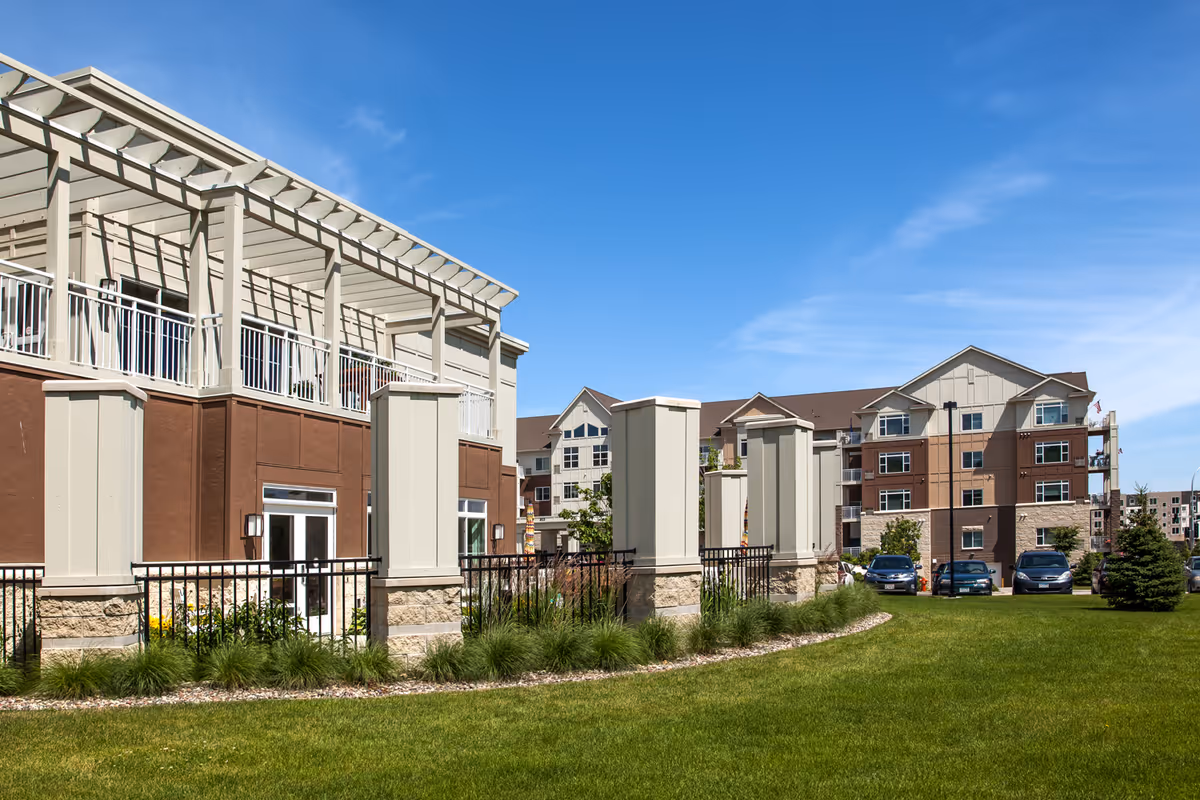 Exterior view of a senior living facility with a well-maintained lawn, decorative pillars, and a multi-story building under a clear blue sky.