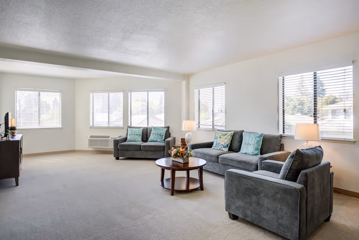 A bright living room with three gray upholstered seating pieces including a sofa, loveseat, and armchair arranged around a round wooden coffee table with a floral centerpiece. The room has multiple windows with white blinds allowing natural light to fill the space. There are two table lamps on either side of the sofa, and a TV on a stand is visible on the left side of the room.