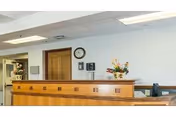 Reception area inside Cliveden Nursing And Rehabilitation Center with a wooden front desk, a wall clock, and floral arrangements on the counter and in the background.