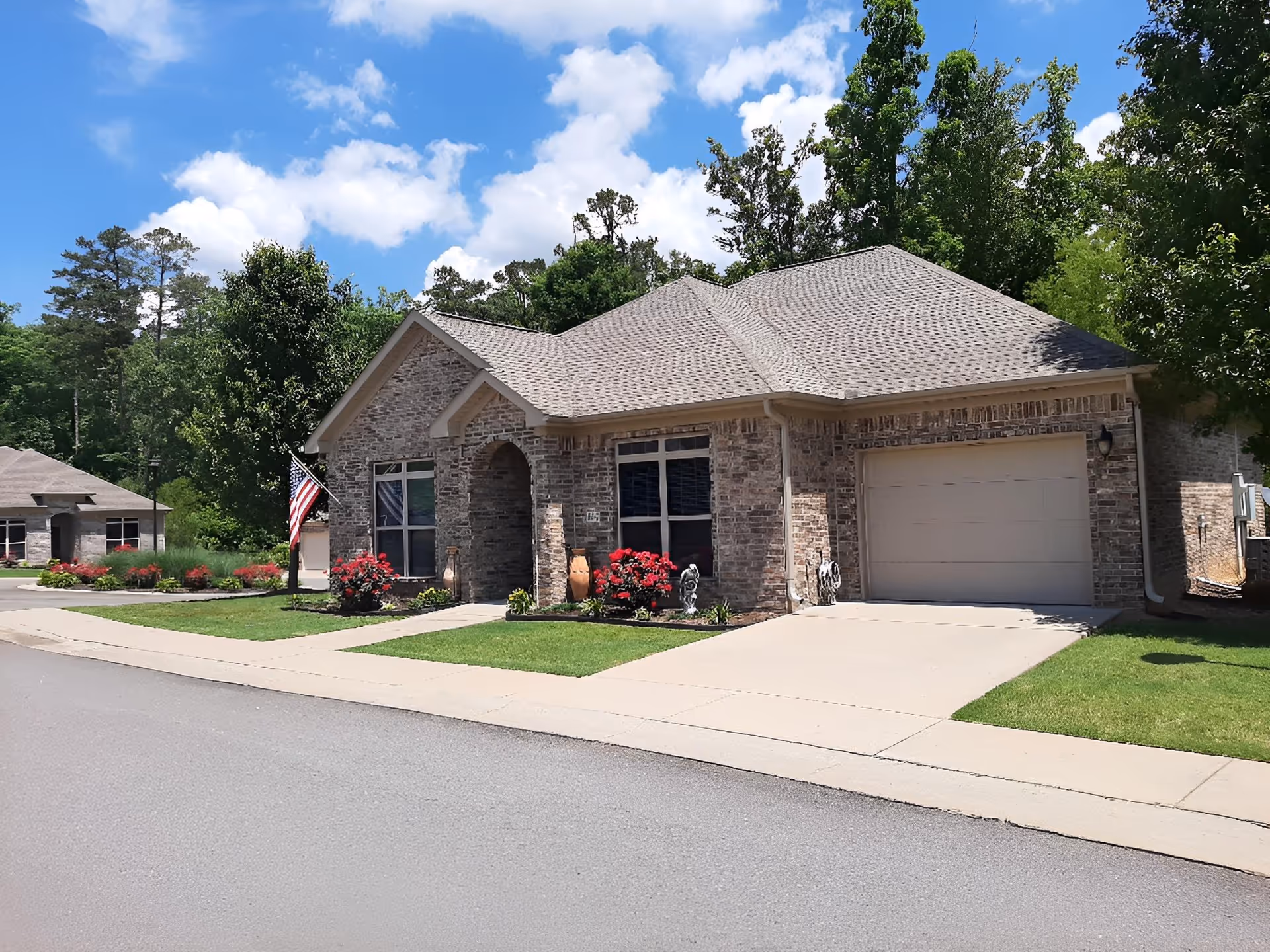 Exterior view of a single-story brick residential building with a garage, a small front lawn with flowers and shrubs, an American flag near the entrance, and a clear blue sky with some clouds.