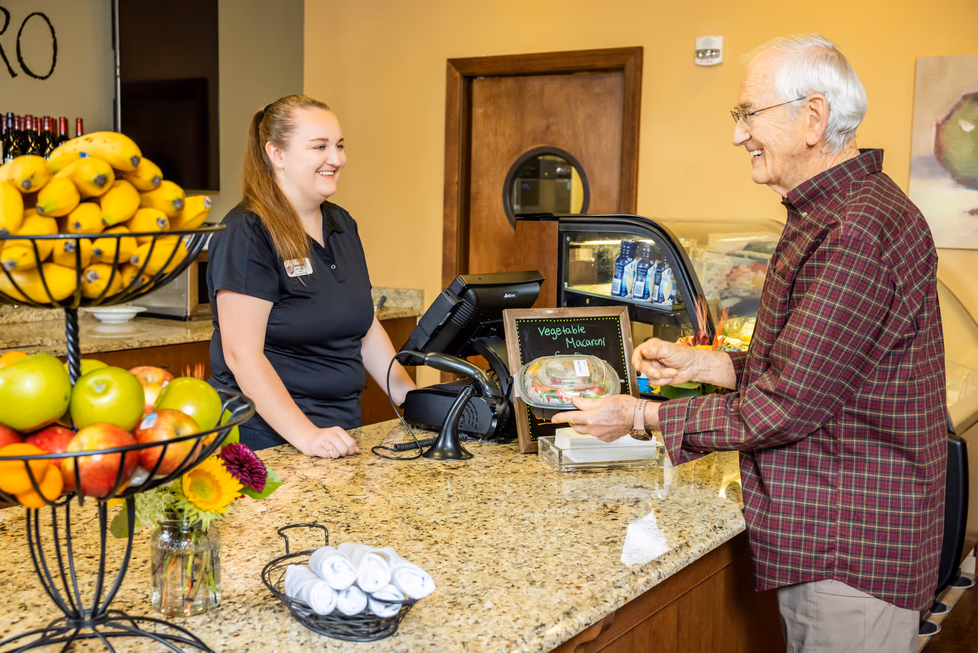 A smiling female staff member stands behind a counter serving an elderly man who is holding a packaged meal labeled 'Vegetable Macaroni.' The counter has a tiered fruit basket with bananas, apples, and oranges, a small vase with flowers, and rolled white napkins. The setting appears to be a food service or dining area with warm yellow walls and a wooden door in the background.