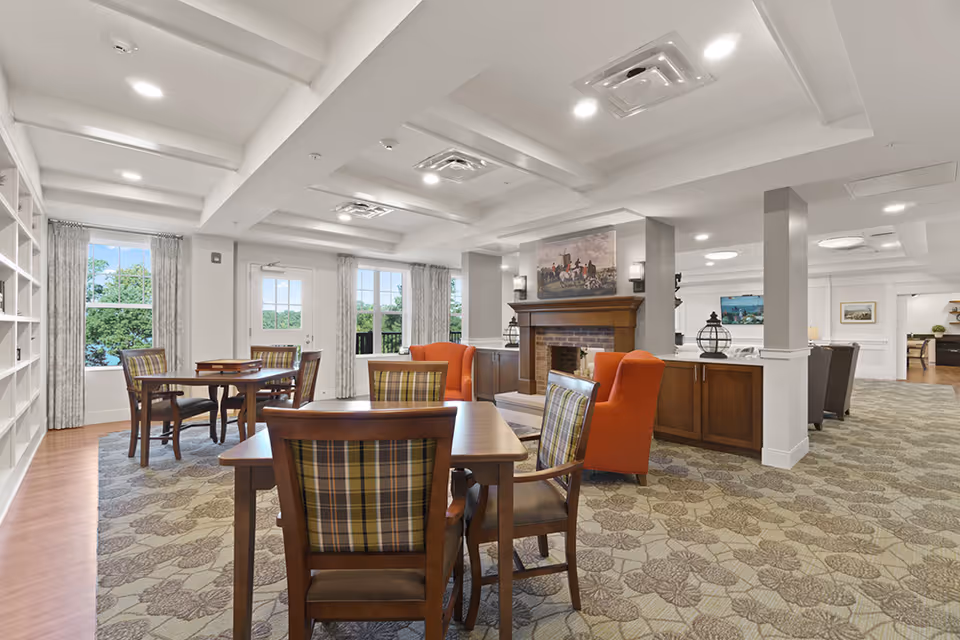 Bright senior living common room with tables and chairs, patterned carpet, an orange armchair by a fireplace, and built-in bookshelves by windows.