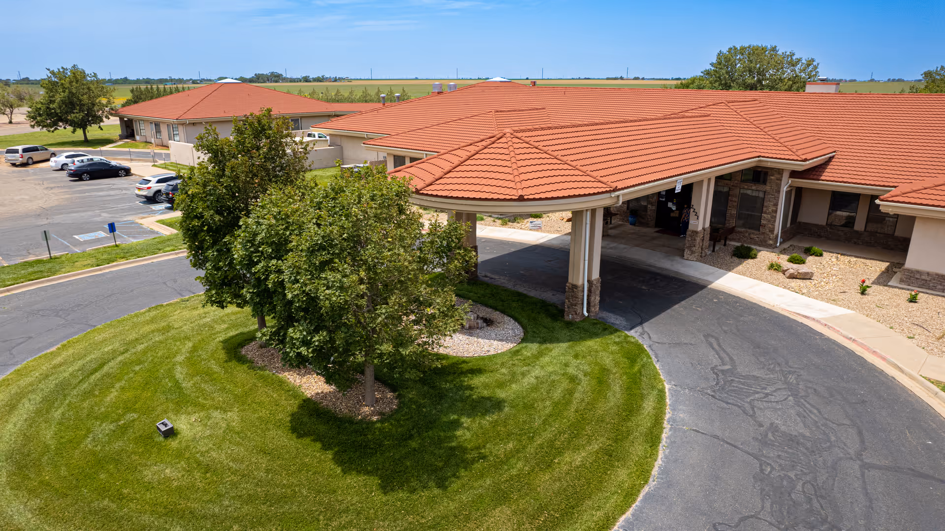Aerial view of the entrance to a single-story senior living facility with a red-tiled roof, a circular driveway, and a landscaped area with green grass and several trees. Several parked cars are visible in the parking lot nearby under a clear blue sky.