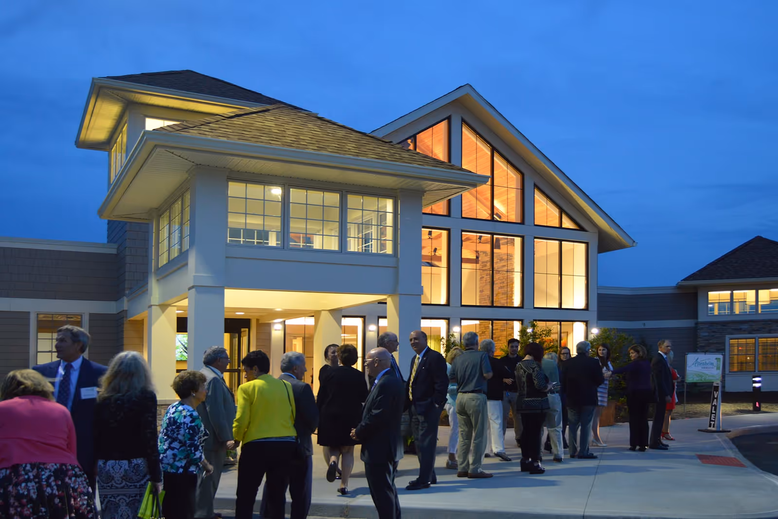 A group of people gathered outside a well-lit senior living facility building during dusk. The building has large windows and a modern design with a peaked roof. Some people are engaged in conversation near the entrance, and a valet sign is visible on the right side.