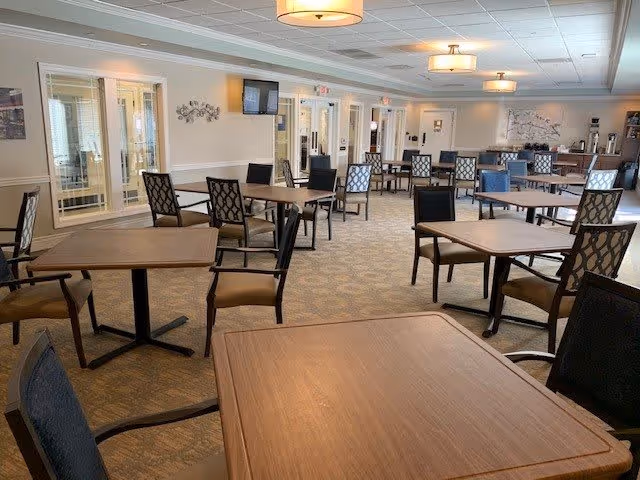 A spacious dining room with multiple wooden tables and cushioned chairs arranged neatly. The room has beige walls, carpeted floors, ceiling lights, and a wall-mounted television. There are glass doors and windows along one side, and a decorative tree painting on the far wall.