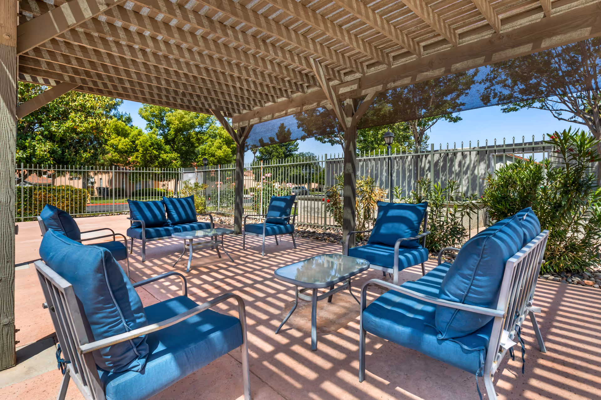 Outdoor seating area under a wooden pergola with blue cushioned chairs and small glass-top tables, surrounded by greenery and a metal fence.