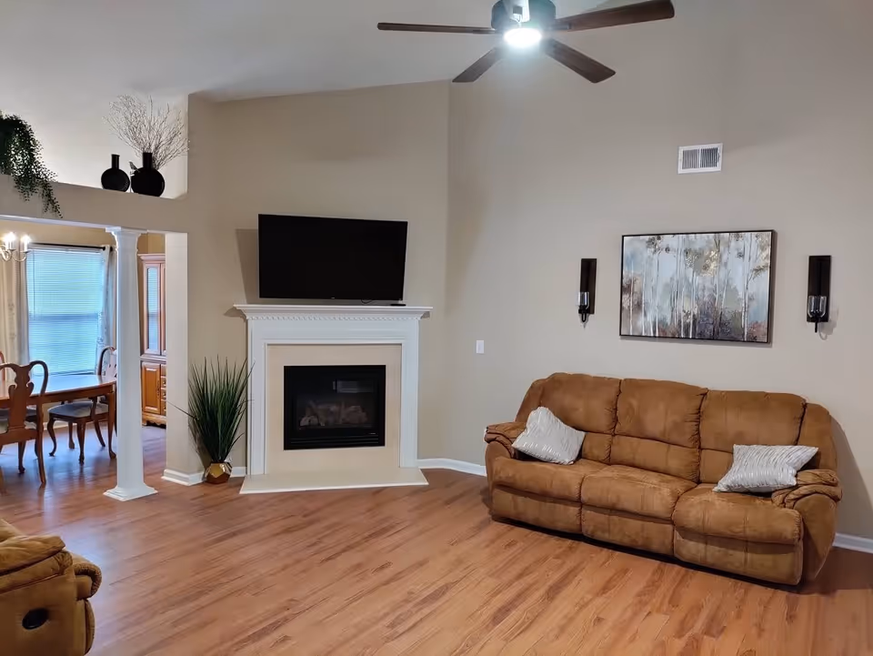 A cozy living room with a brown three-seater sofa adorned with two silver pillows. A white fireplace with a black TV mounted above it is centered on the far wall. To the left, there is an open entryway with white columns leading to a dining area with wooden chairs and a table. The room has wooden flooring, beige walls, a ceiling fan with a light, and decorative wall sconces flanking a framed painting of trees.