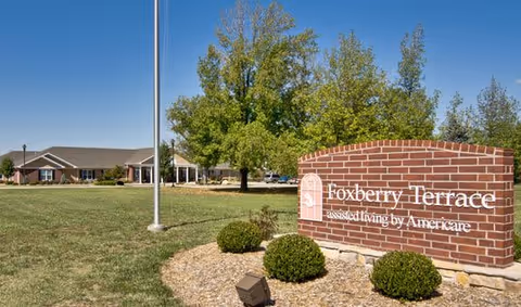 Exterior view of Foxberry Terrace Senior Living facility with a large brick sign in the foreground displaying the facility's name and a single-story building in the background surrounded by grass and trees under a clear blue sky.
