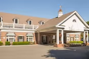 Brick senior living facility front with a covered entrance portico, white columns, a balcony, and a driveway under a clear blue sky.