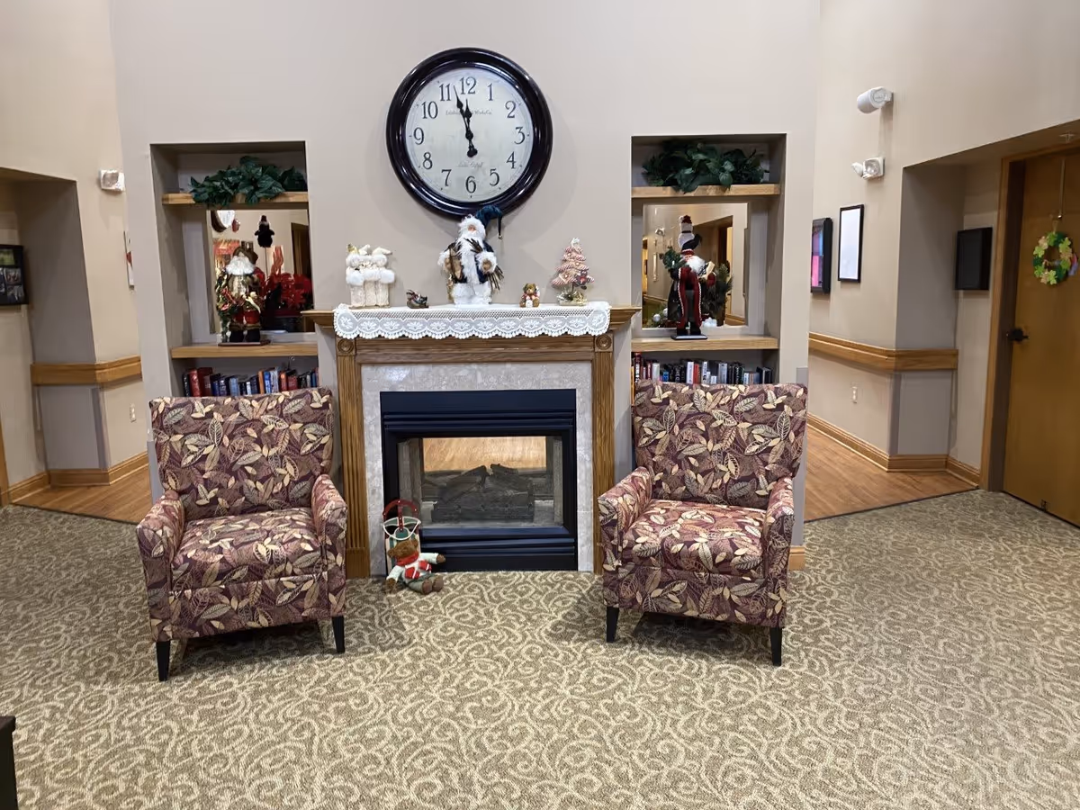 Cozy common room with a central fireplace, a large wall clock, two patterned armchairs, bookshelves and holiday decorations.