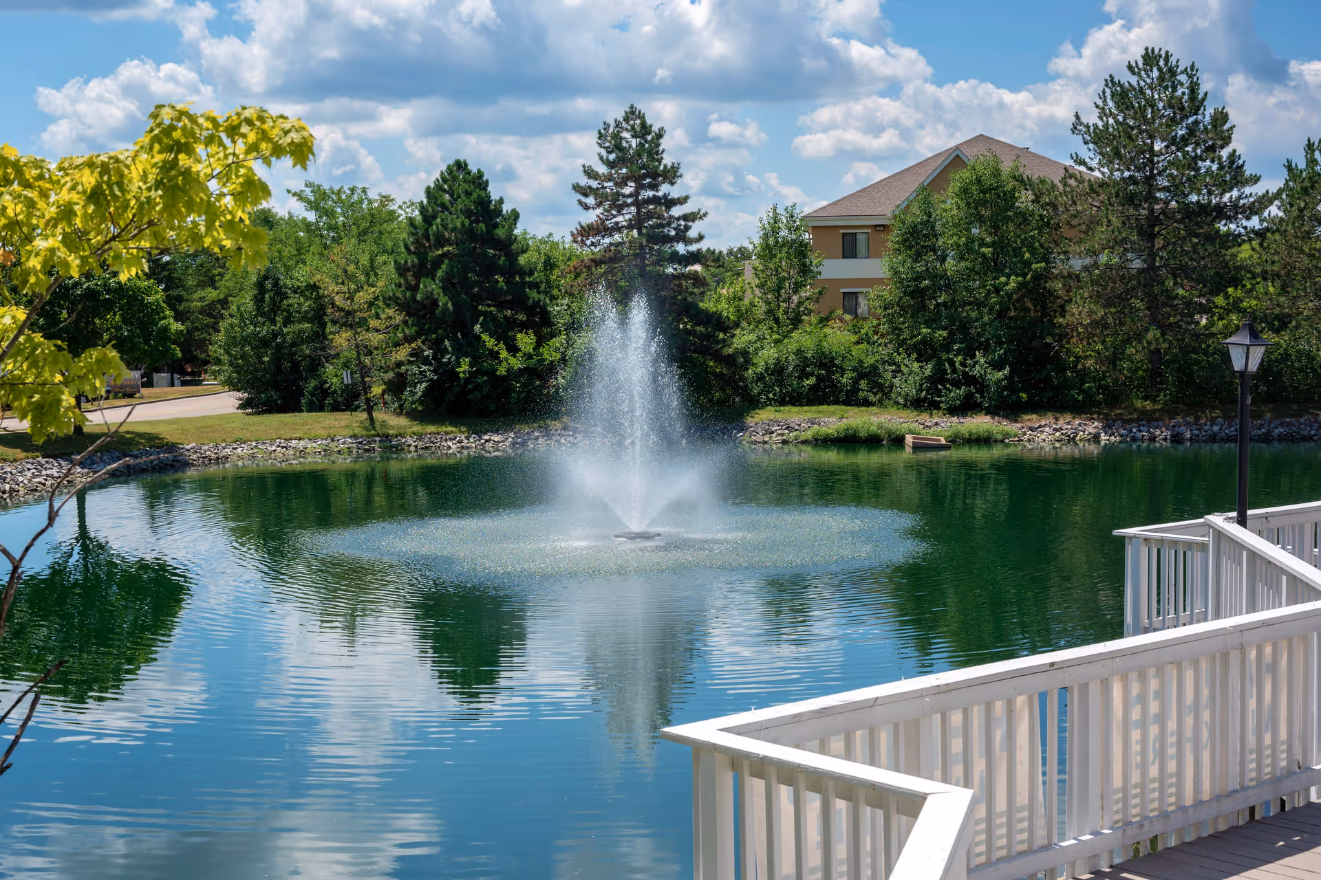 A peaceful outdoor scene featuring a pond with a water fountain in the center, surrounded by green trees and shrubs. A beige building with a brown roof is visible in the background. In the foreground, there is a white wooden railing and a black lamp post on a deck or walkway. The sky is partly cloudy with blue patches.