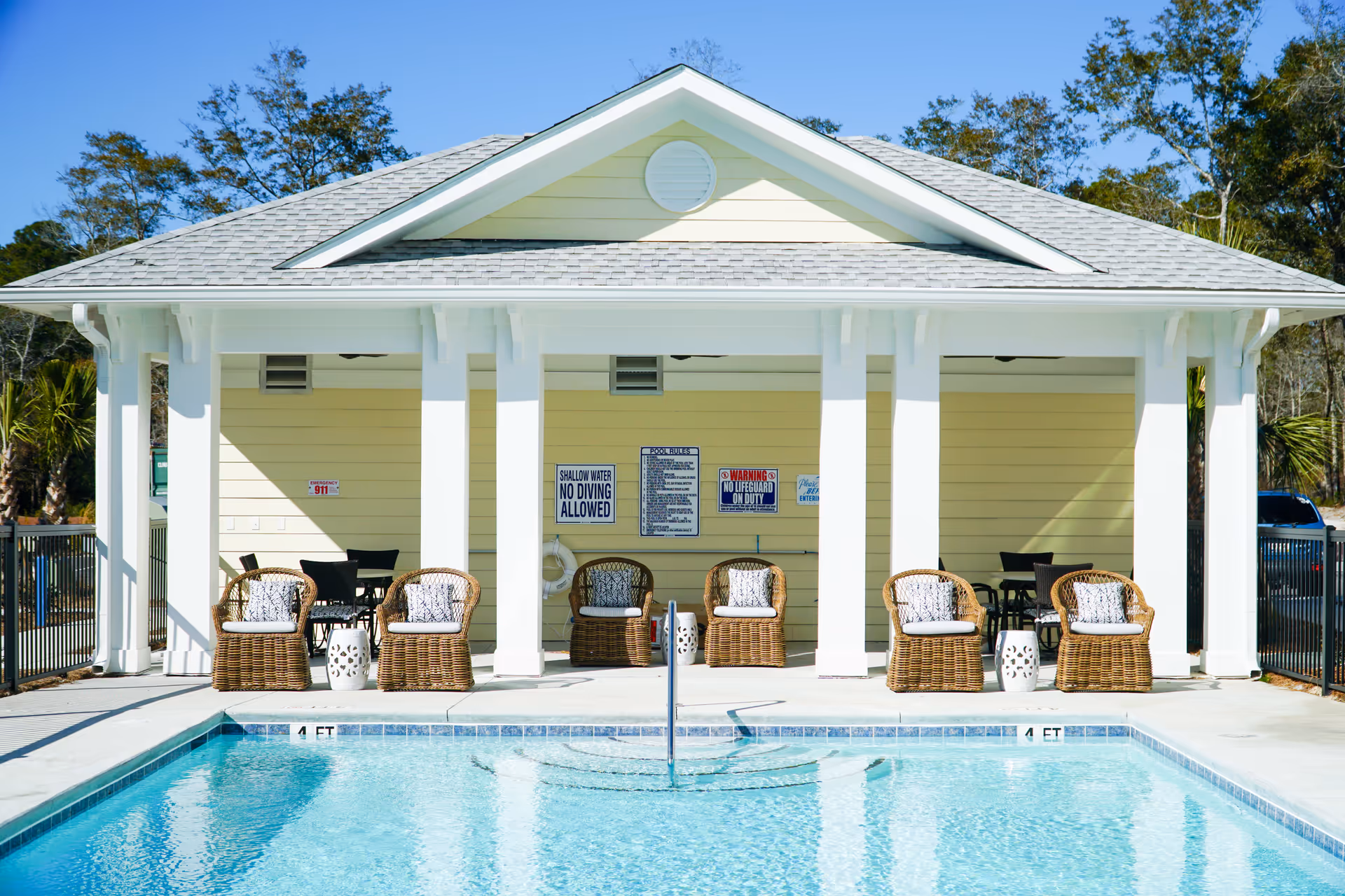 Outdoor swimming pool with clear blue water and steps leading into the pool. Behind the pool is a covered seating area with six wicker chairs, each with a cushion and pillow, and two small white side tables. The building behind the seating area is light yellow with white columns and a gray shingled roof. Several safety and pool rules signs are visible on the wall.