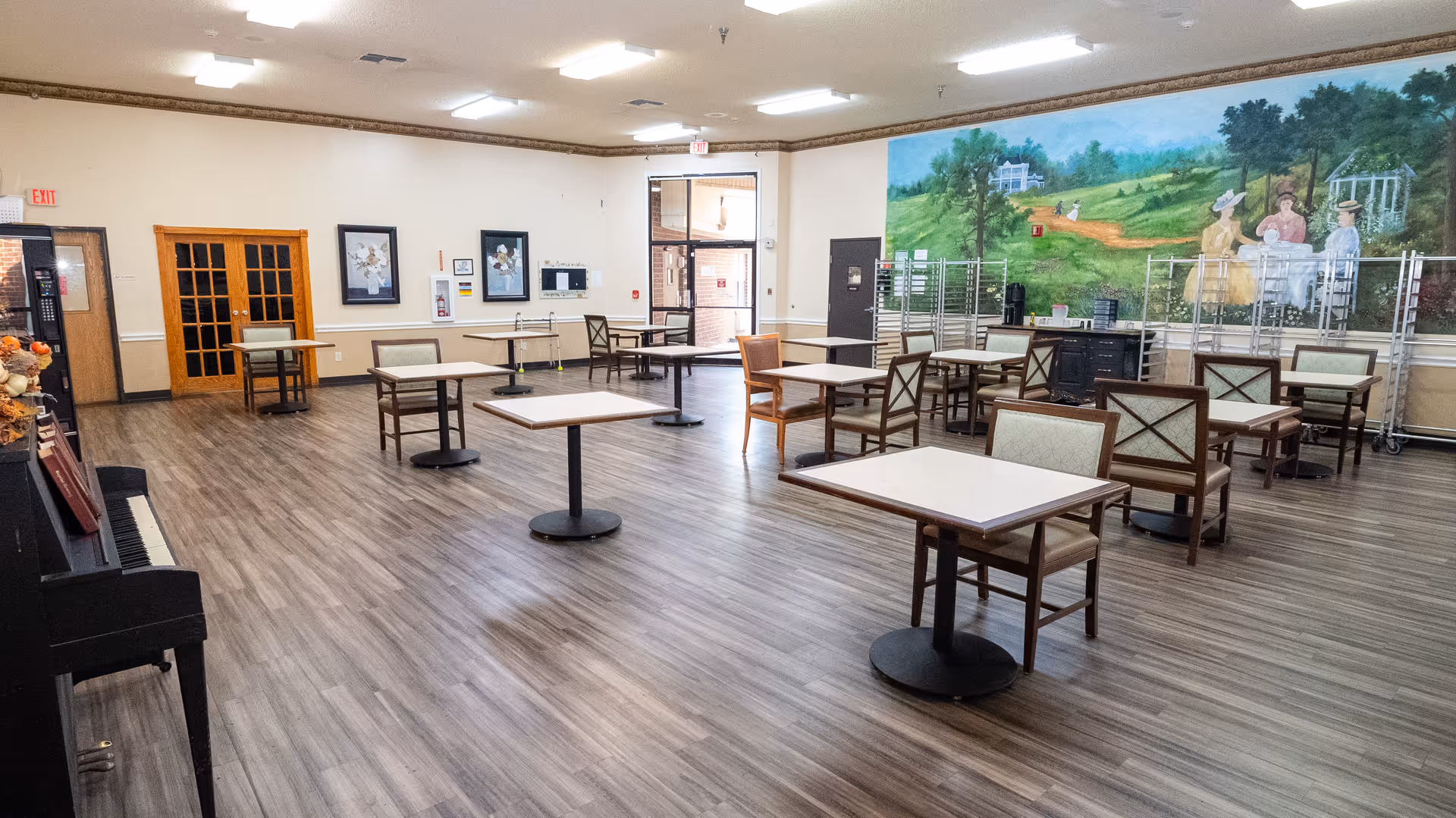 Spacious senior facility dining/activity room with spaced tables and chairs, wood-look flooring and a large mural on the far wall.
