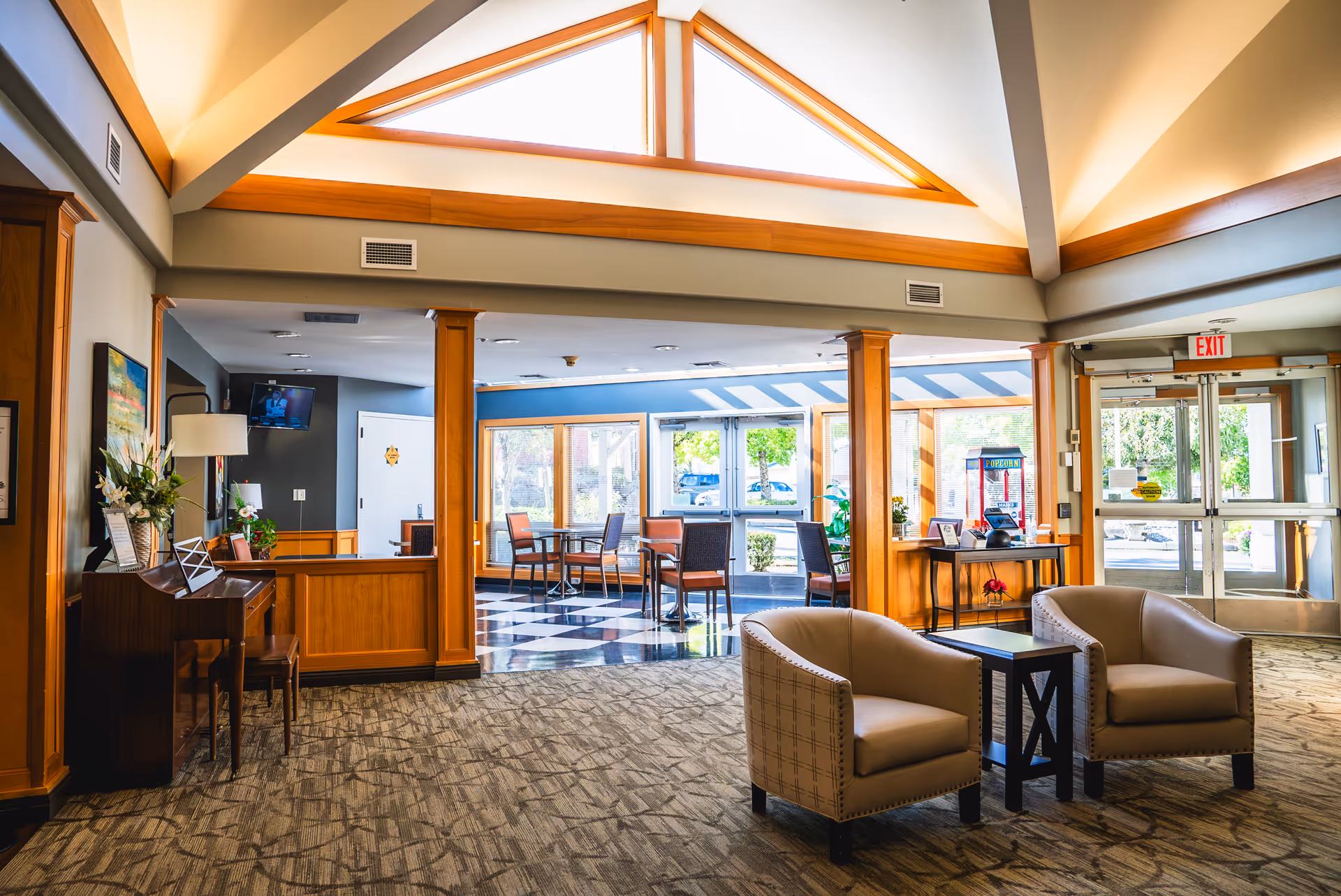 A bright and spacious assisted living facility lobby with high vaulted ceilings and large triangular windows letting in natural light. The area features two beige armchairs with a small black table between them on a patterned carpet. In the background, there is a seating area with tables and chairs near large windows, a piano with a lamp and flowers on it, and an exit door with glass panels.