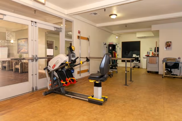 Rehabilitation/therapy room with a seated exercise machine, parallel bars, and other treatment equipment.