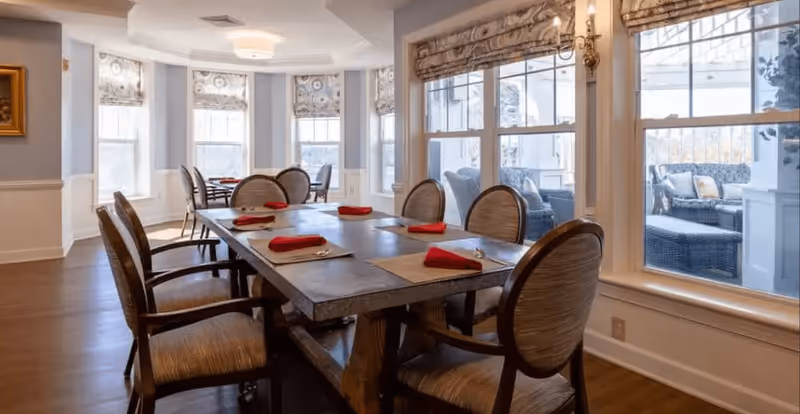 A dining room with a large wooden table set with placemats and red folded napkins. The room has several upholstered chairs around the table and large windows with patterned Roman shades, allowing natural light to fill the space. Outside the windows, there is a view of a patio area with wicker furniture and cushions.