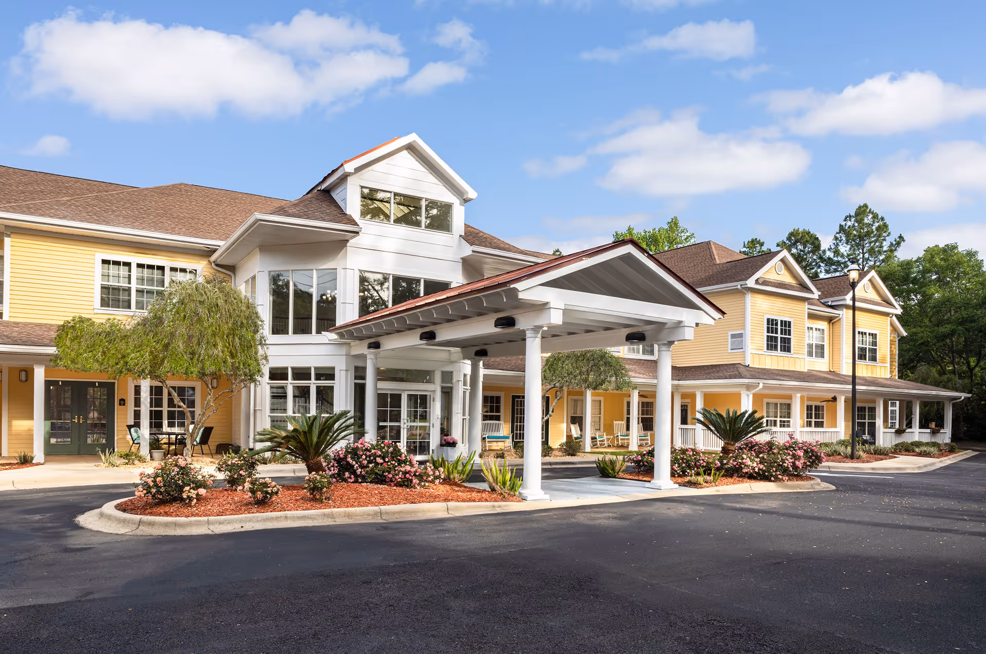 Exterior view of The Gables of Tallahassee senior living facility showing a large yellow building with white trim, a covered entrance supported by white columns, landscaped flower beds with bushes and small trees, and a paved driveway under a partly cloudy sky.