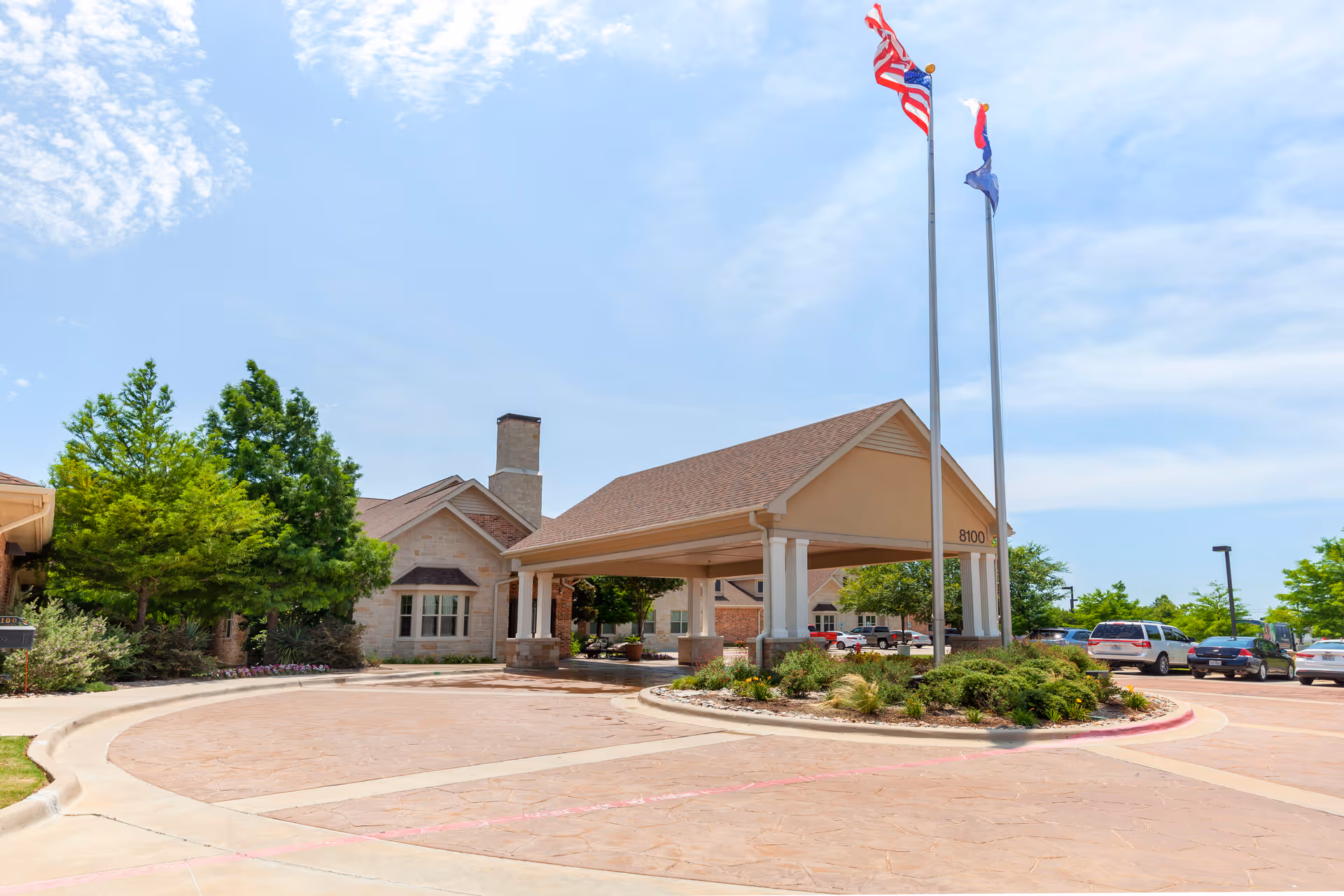 Exterior view of a senior living facility entrance with a covered driveway, two flagpoles with flags, surrounding greenery, and parked cars under a partly cloudy sky.