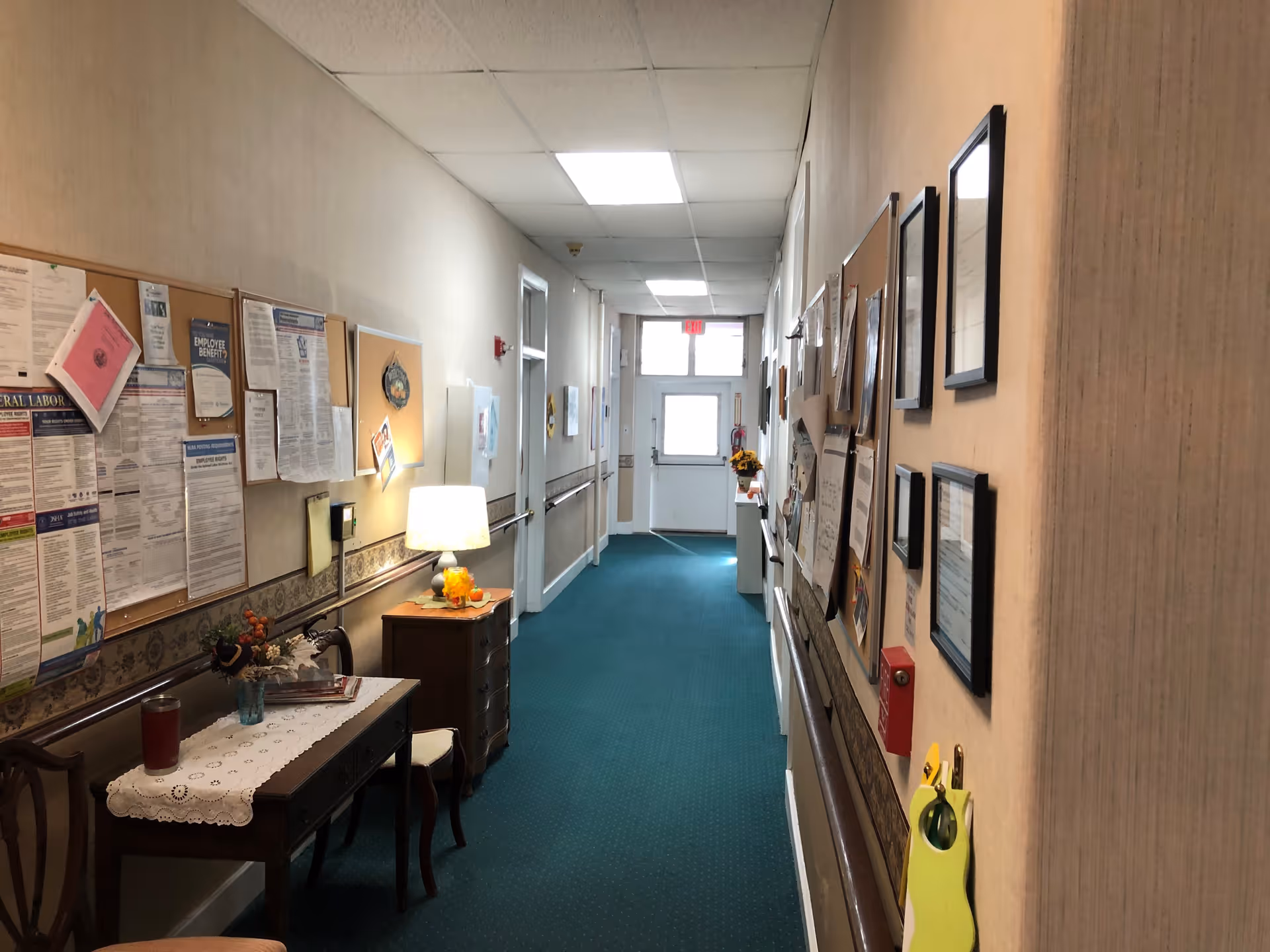 A long, narrow hallway with teal carpet and beige walls decorated with bulletin boards, framed documents, and notices. There is a small table with a lace cloth, a chair, and a lamp on a wooden dresser along the left side. The hallway is well-lit with ceiling lights and natural light coming from a door with windows at the far end.