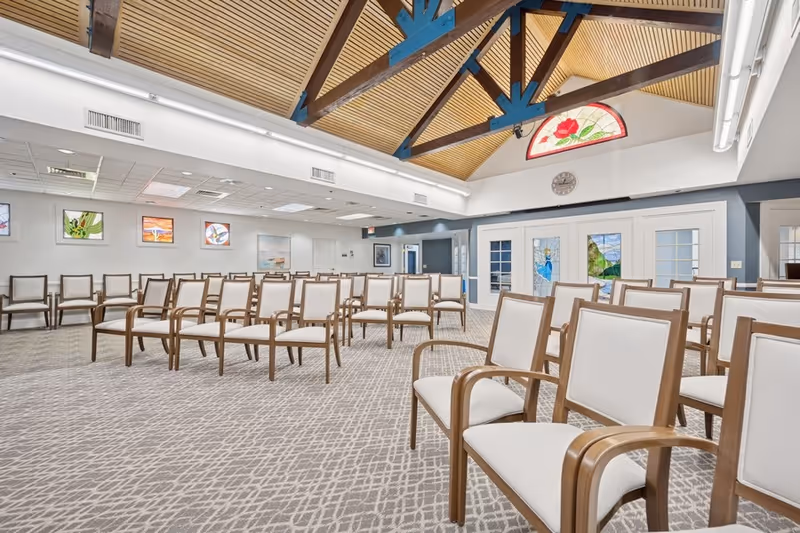 A spacious room with rows of wooden chairs with white cushions arranged facing forward. The room has a high ceiling with wooden paneling and blue beams, decorative stained glass windows, and framed artwork on the walls. The carpet has a geometric pattern, and the space appears well-lit with fluorescent lighting.