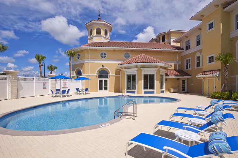 Outdoor swimming pool area at The Terraces at Bonita Springs with blue lounge chairs and umbrellas around the pool. The building in the background has a yellow exterior with a red-tiled roof and a small tower structure. Palm trees and a bright blue sky with some clouds are visible.