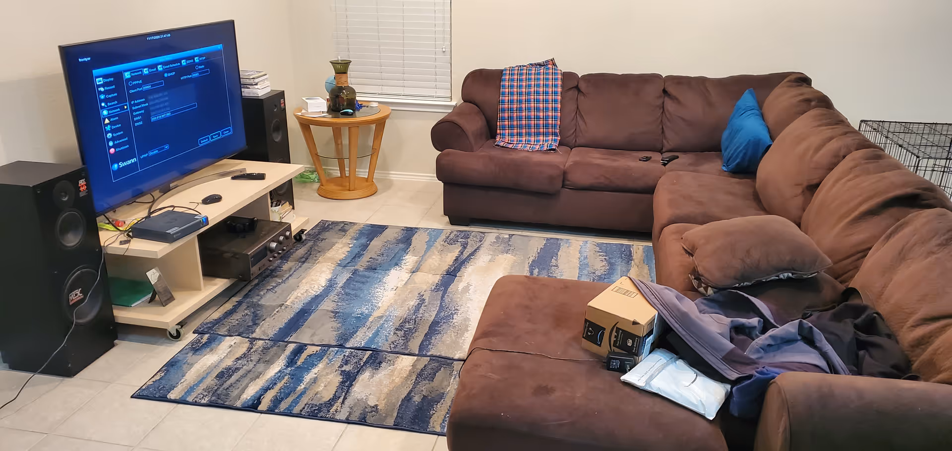 A living room with a large brown sectional sofa, a blue and beige patterned rug, a TV on a light wood stand with speakers on each side, and a small round wooden side table with decorative items. There is a window with closed blinds behind the sofa.