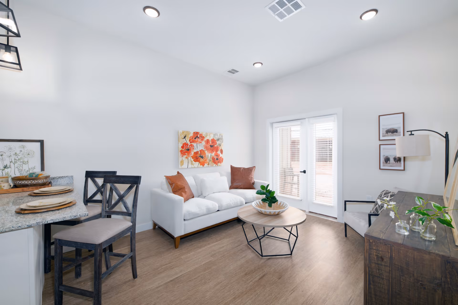 A bright and modern living room with a white sofa adorned with two brown pillows, a round wooden coffee table with a plant centerpiece, two dark wooden chairs at a granite countertop, and a wooden sideboard with decorative items. The room has light wood flooring, white walls, recessed ceiling lights, and French doors with blinds leading outside. Artwork featuring red flowers and animal prints decorate the walls.