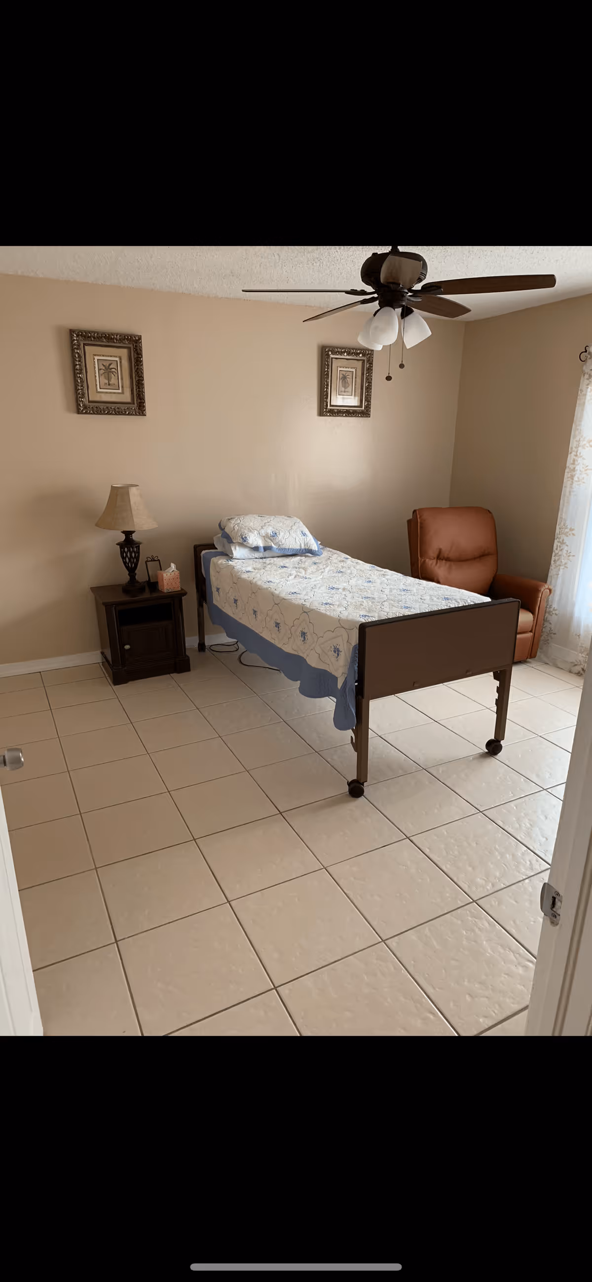 A simple bedroom with a single bed covered with a white and blue patterned quilt, a wooden nightstand with a lamp and tissue box, a brown recliner chair near a window with sheer curtains, two framed pictures on the beige wall, and a ceiling fan with lights.