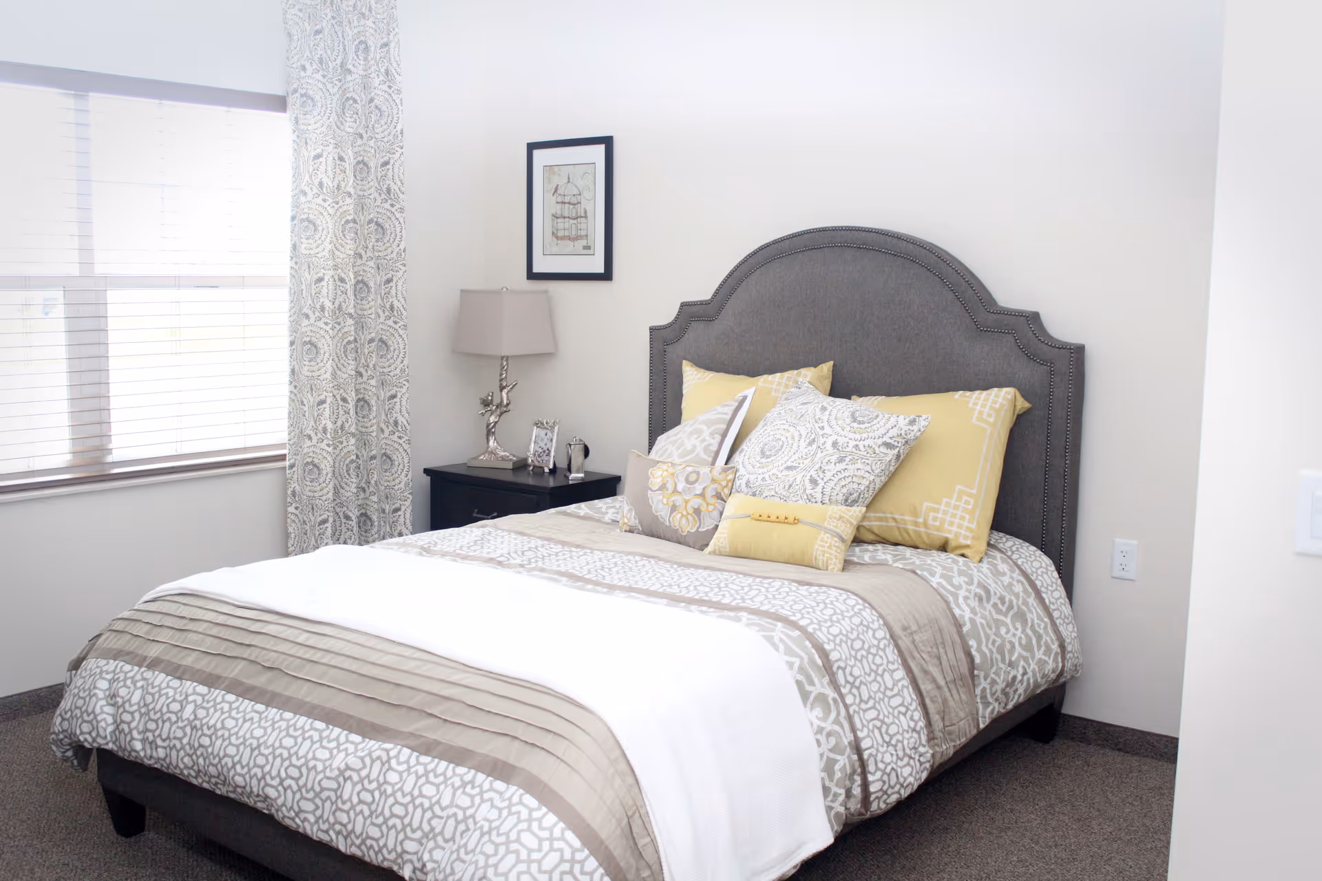 Bright bedroom with a neatly made bed featuring patterned pillows and a gray upholstered headboard, a nightstand with a lamp, and a window with patterned curtains.