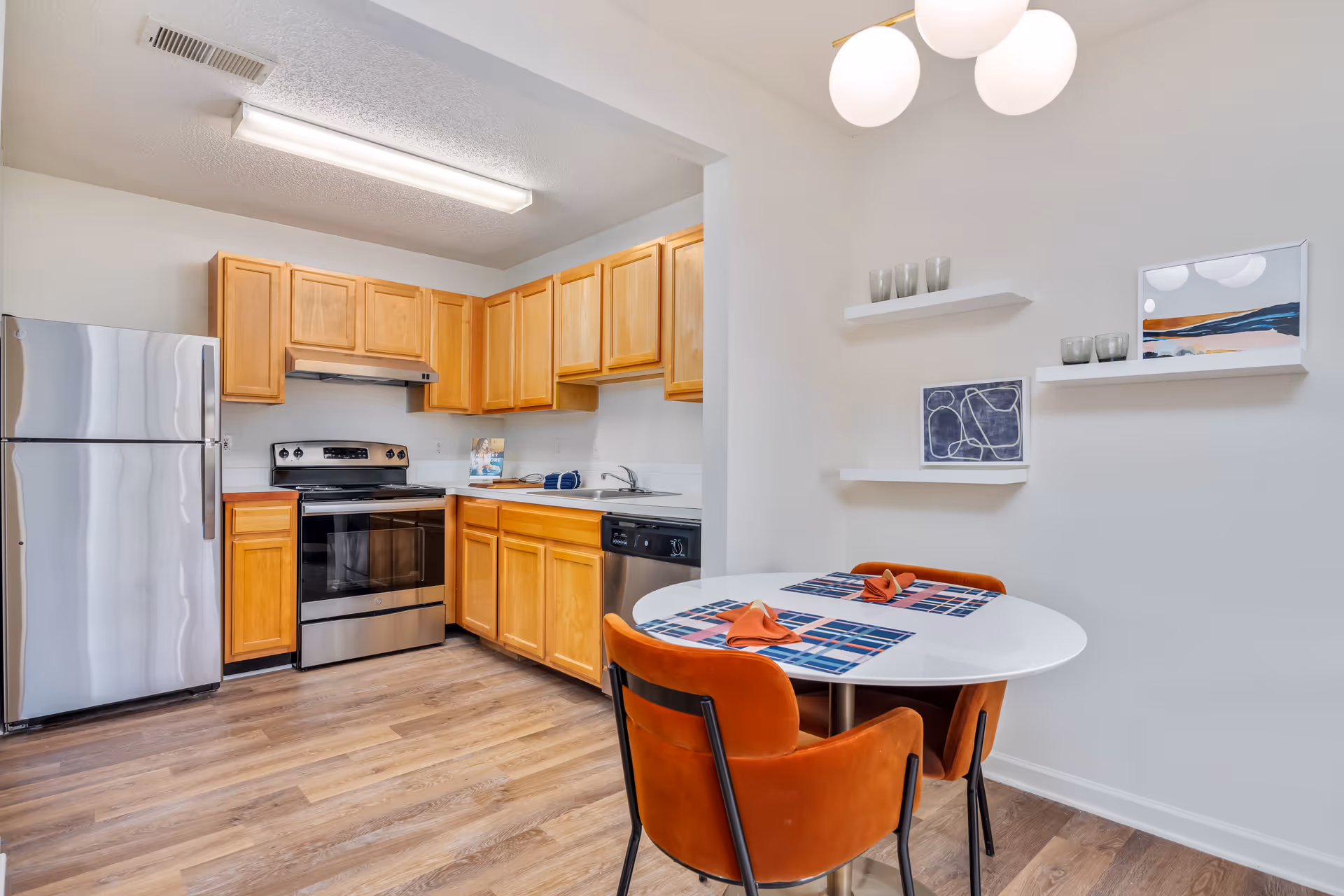 A modern kitchen and dining area featuring wooden cabinets, stainless steel refrigerator, oven, and dishwasher. The dining area has a round white table with two orange upholstered chairs, blue and orange placemats, and folded orange napkins. The wall has three white floating shelves with decorative glassware and framed artwork. The floor is wood laminate, and the ceiling has a fluorescent light in the kitchen and a modern light fixture above the dining table.