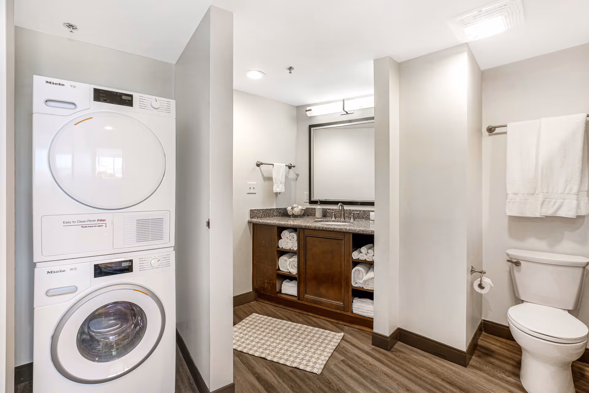 A modern bathroom featuring a stacked washer and dryer, a vanity with granite countertop and open shelving of towels, a toilet, and towel racks.