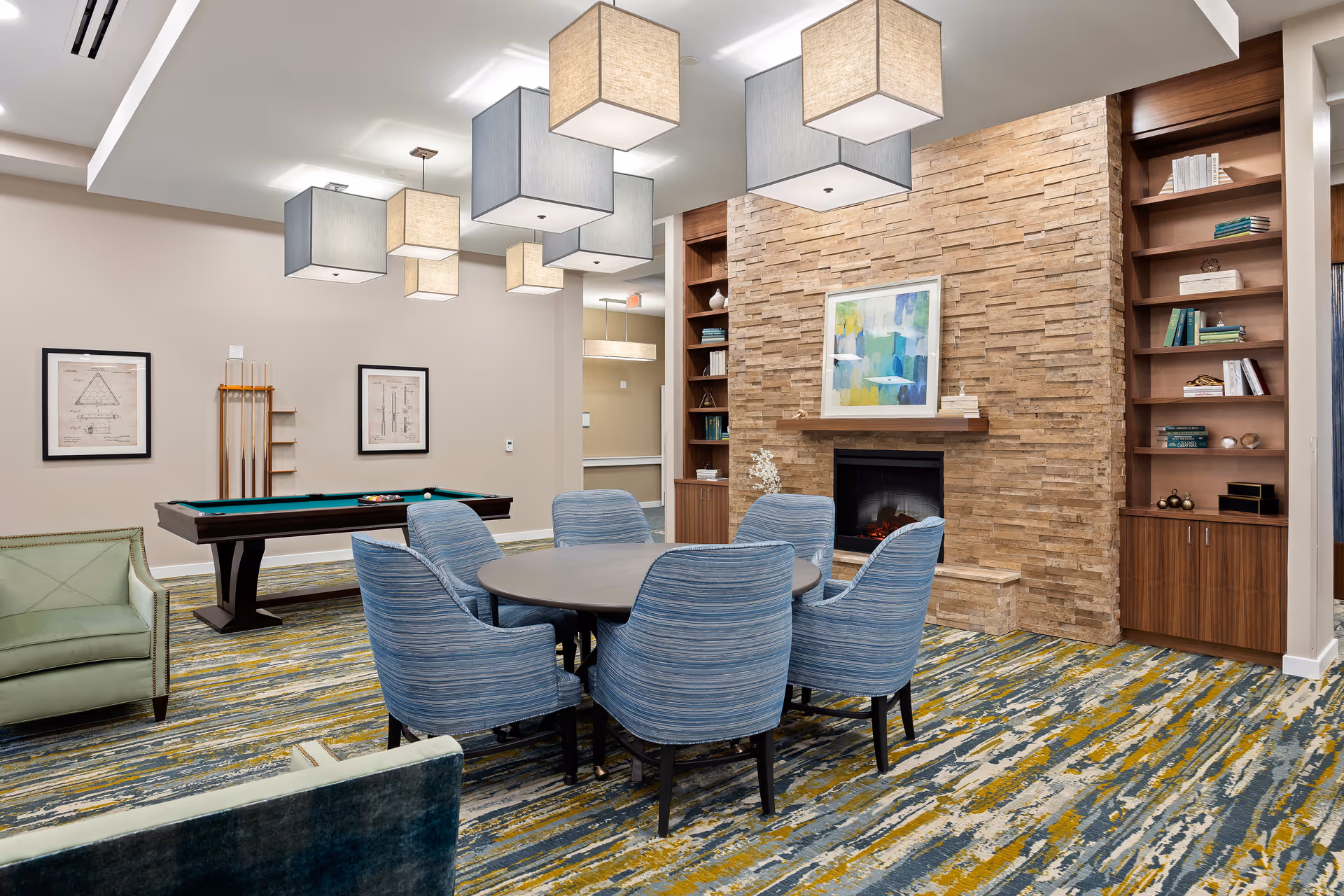 A cozy common area featuring a round table surrounded by six blue upholstered chairs, a stone fireplace with a colorful abstract painting above it, built-in wooden bookshelves filled with books and decorative items, a pool table with cues on the wall, and modern square pendant lights hanging from the ceiling. The room has a patterned carpet with blue, yellow, and gray tones.