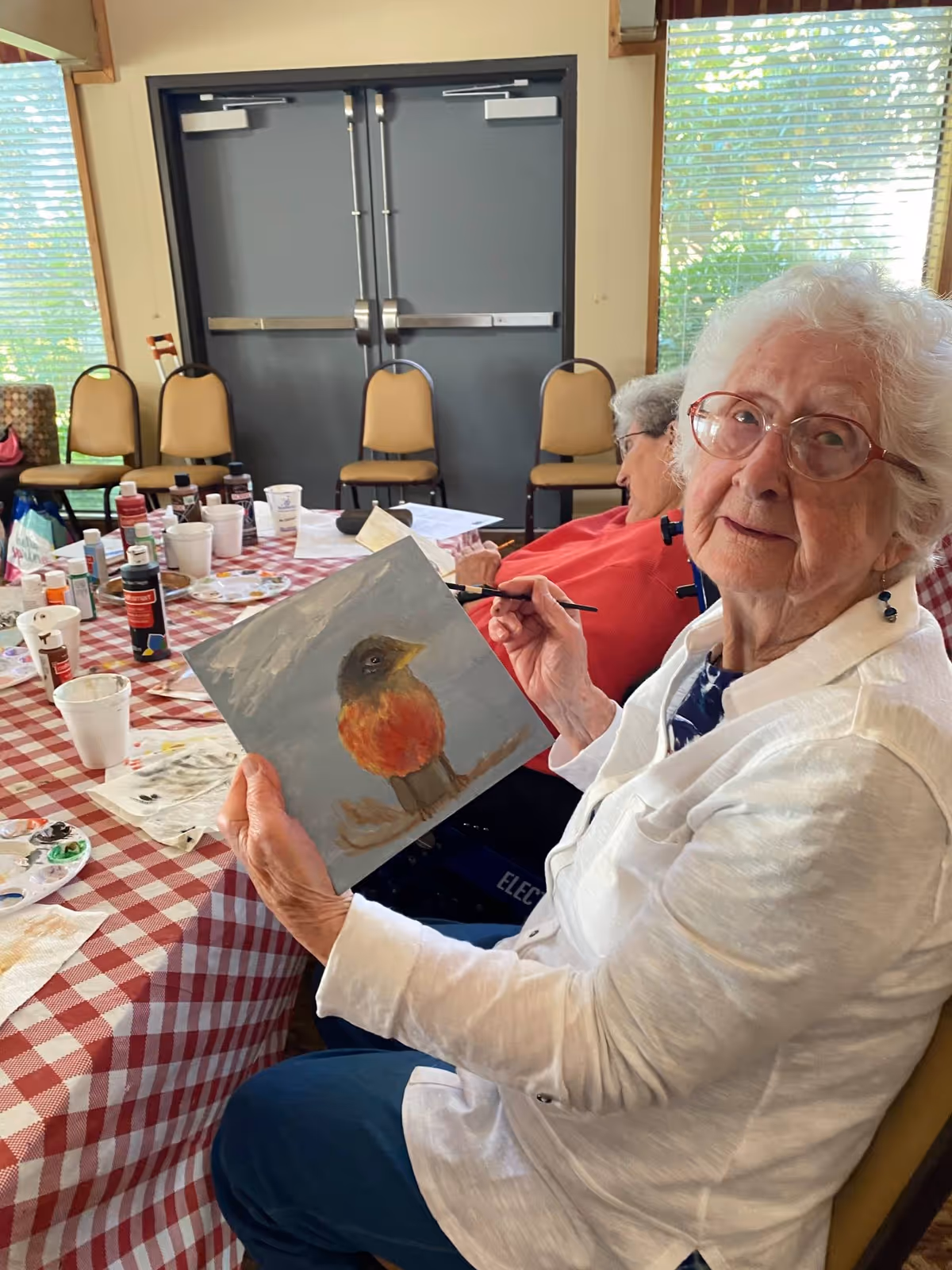 An elderly woman with white hair and glasses is sitting at a table covered with a red and white checkered tablecloth, holding a painting of a bird with an orange chest. She is painting with a brush and looking at the camera. Another elderly person is seated beside her, also engaged in painting. The room has large windows with blinds and several chairs against the wall.