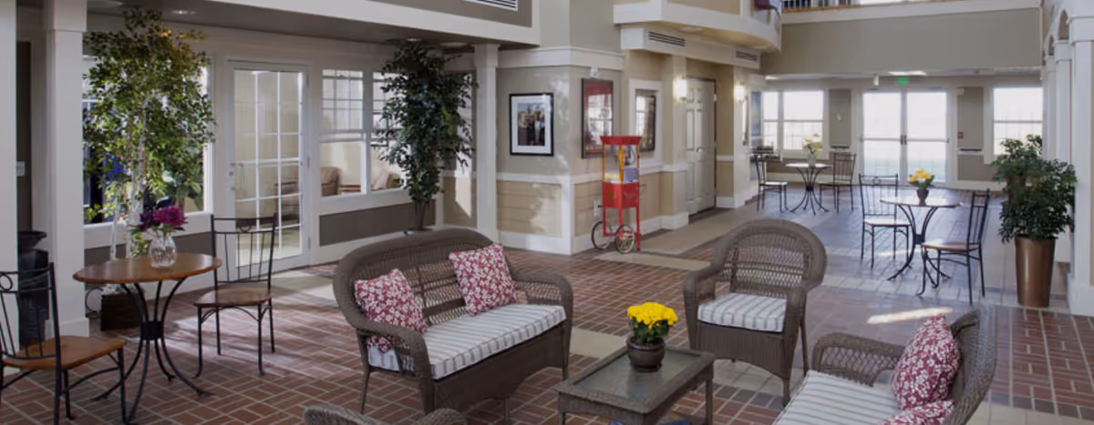 A spacious indoor common area with brick flooring, wicker seating with striped cushions and floral pillows, small round tables with chairs, potted plants, and large windows letting in natural light.